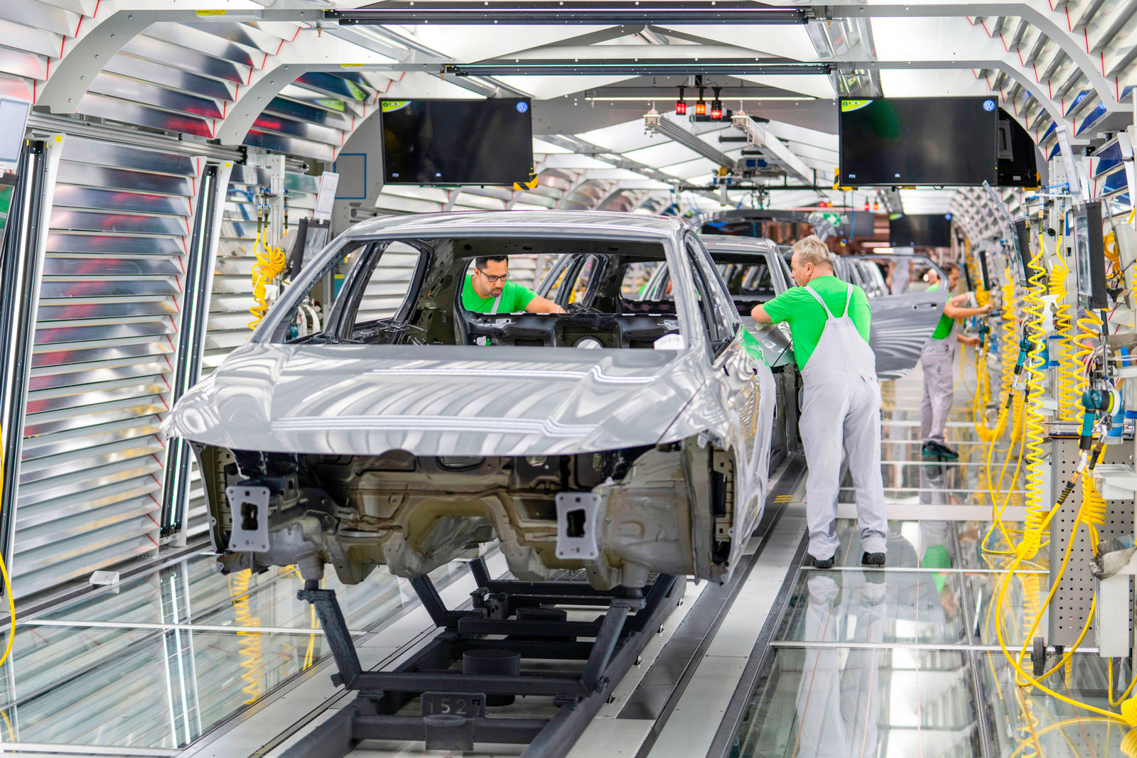 A modern automotive assembly line with partially assembled car bodies on a conveyor belt. Workers in green shirts and white overalls are installing components and inspecting quality. The environment is brightly lit with reflective metal surfaces and yellow pneumatic hoses on the sides.