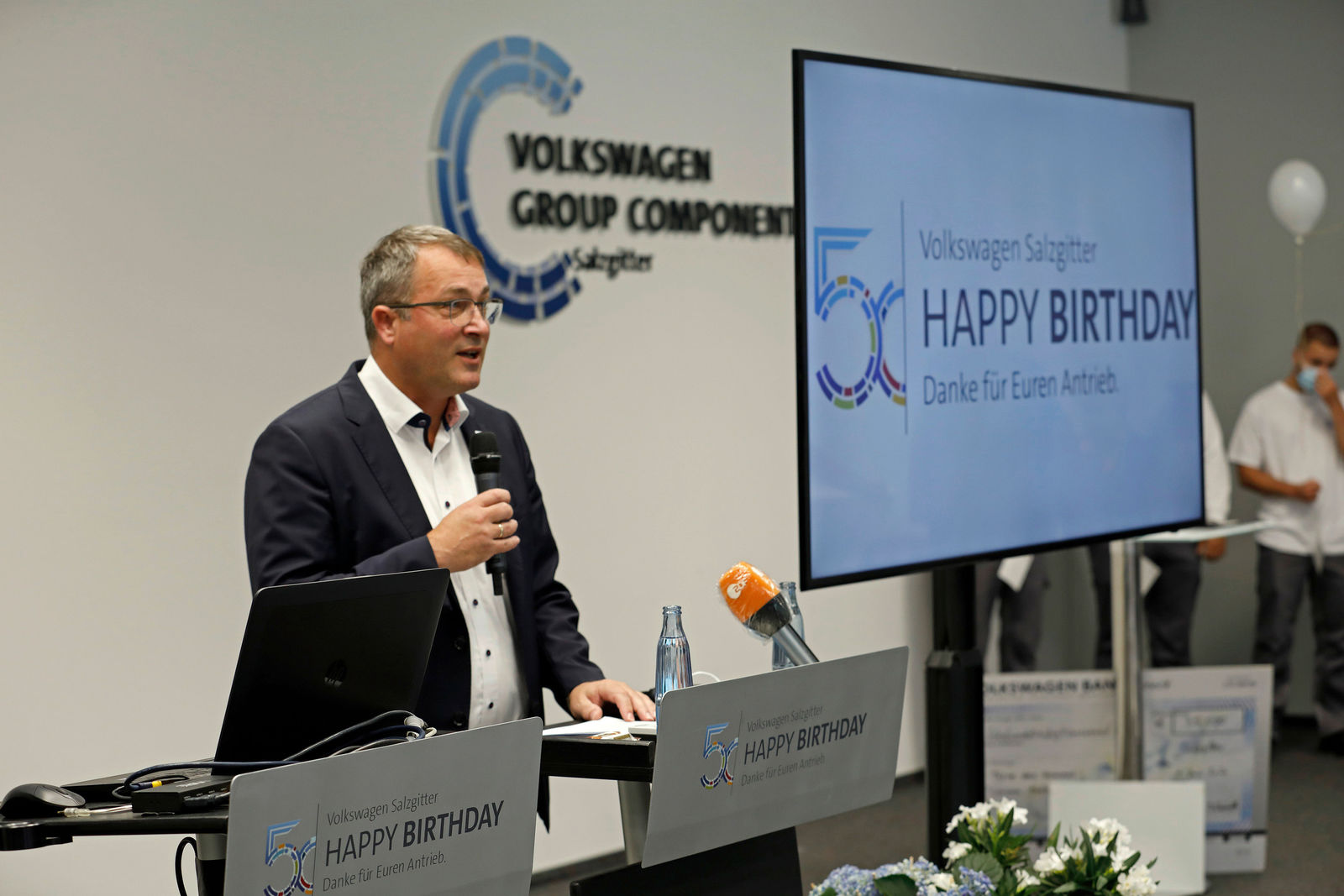 Ein Mann spricht am Rednerpult in einem Raum, der für eine Feier dekoriert ist. Auf dem Rednerpult steht "Volkswagen Salzgitter - Happy Birthday - Danke für Euren Antrieb," und auf einem großen Bildschirm im Hintergrund ist das gleiche Logo zu sehen, das den 50. Geburtstag der Einrichtung feiert. Das Logo der Volkswagen Group Components ist an der Wand zu erkennen.