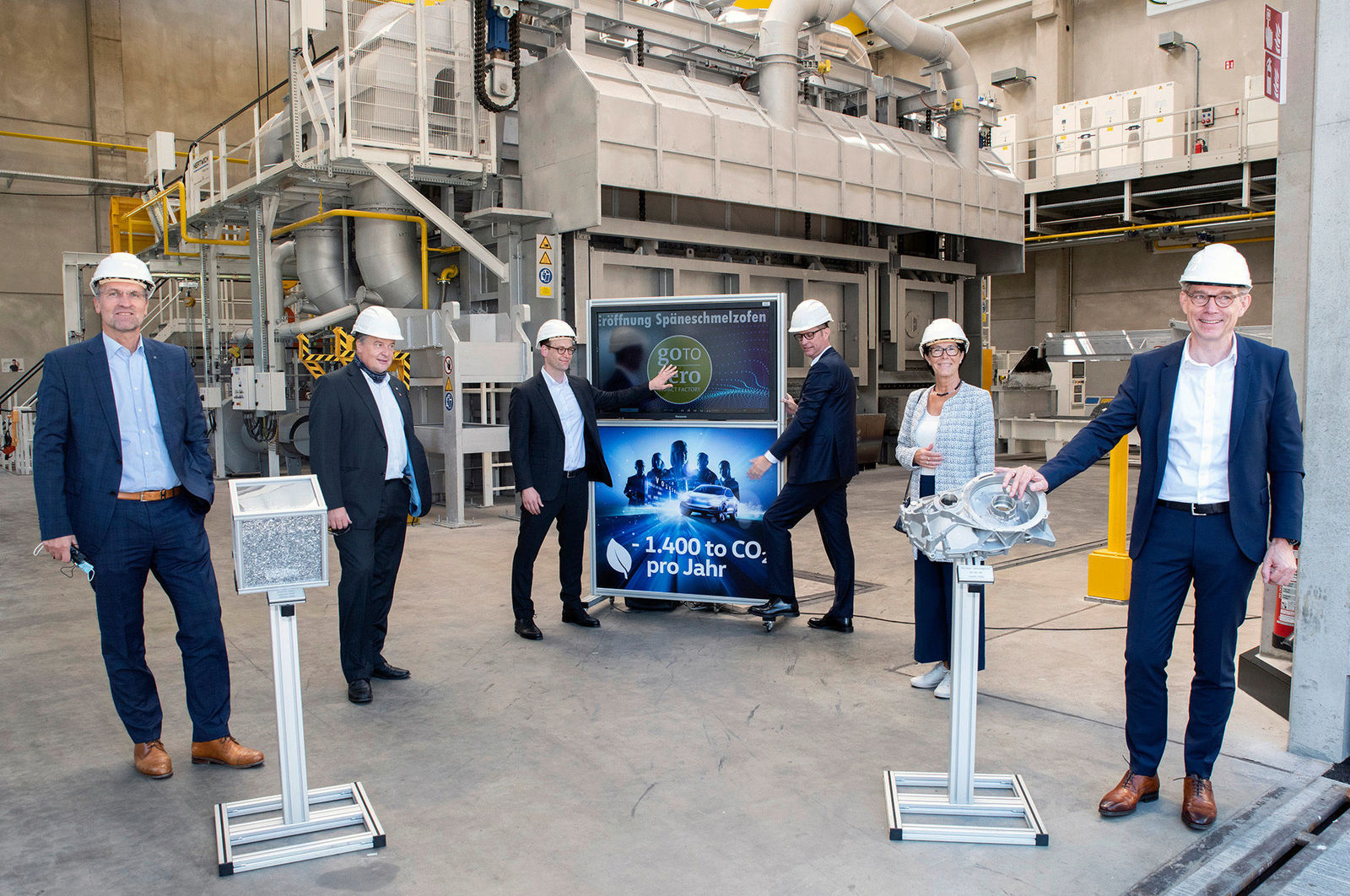 Eine Gruppe von Personen in Schutzhelmen steht in einer industriellen Umgebung vor einem neuen Schmelzofen. In der Mitte wird ein Bildschirm gezeigt, der den Text „1.400 t CO₂ pro Jahr“ und das Logo „goTOzero“ enthält. Die Szene hebt die Eröffnung eines Spaneschmelzofens hervor, der zur Reduktion von CO₂-Emissionen beiträgt.