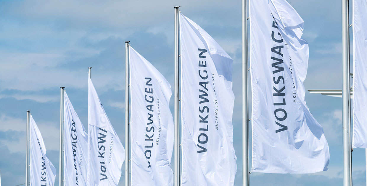 Seven flagpoles can be seen. The white flags bear the dark blue inscription “Volkswagen - Aktiengesellschaft”. The flags rise into the light blue, slightly cloudy sky.