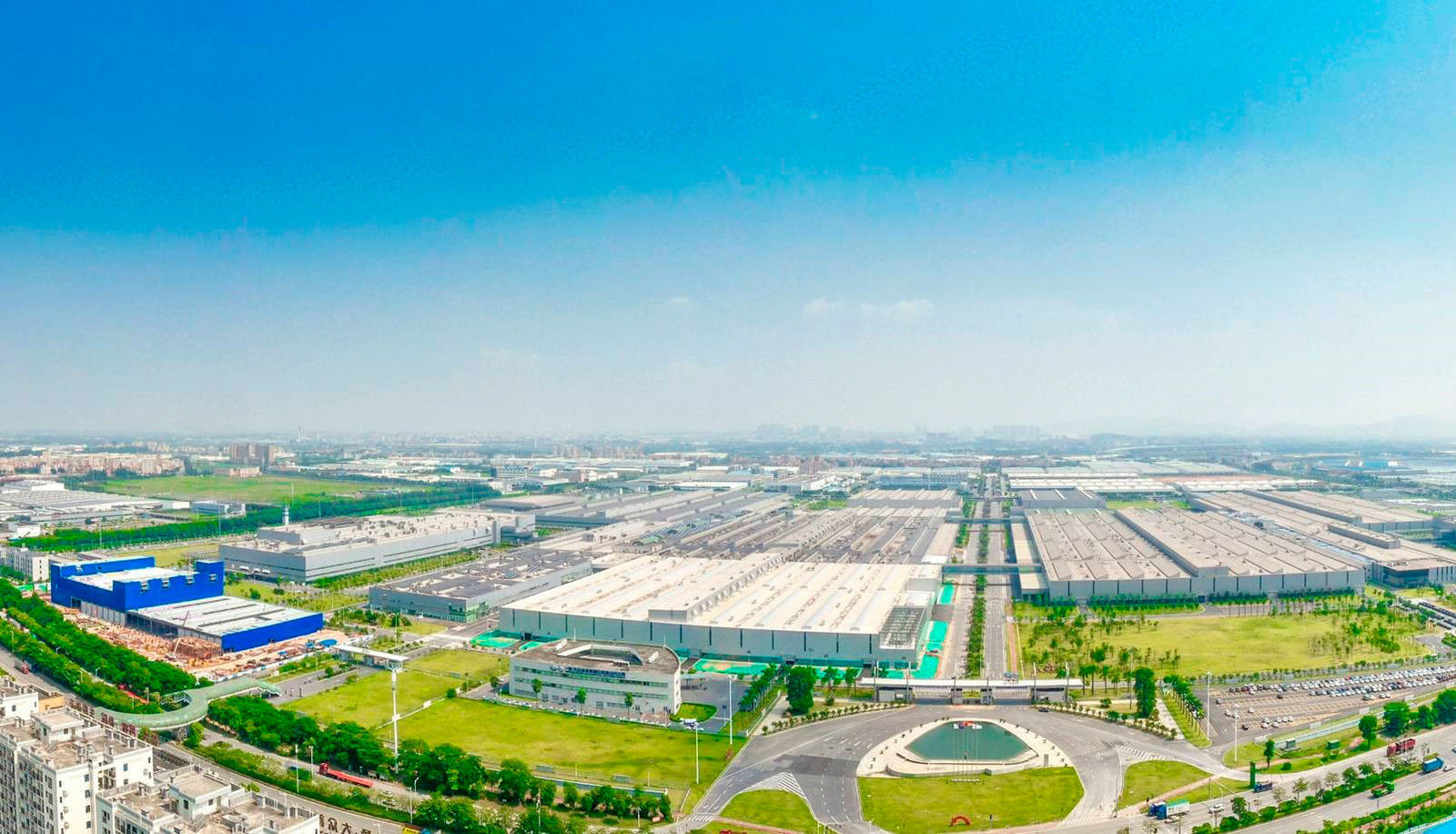 Aerial view of a large industrial complex with multiple extensive halls and buildings. In the foreground, there is a circular plaza with a water feature in the center. The factory site is surrounded by green areas and roads, extending far into the landscape.