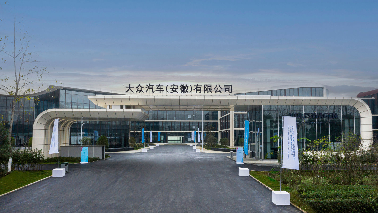 A modern building with a large glass facade featuring the Chinese inscription "大众汽车（安徽）有限公司," which translates to "Volkswagen (Anhui) Co., Ltd." The entrance area is designed with a wide pathway and decorative plants, flanked by banners on both sides. The sky is slightly overcast.