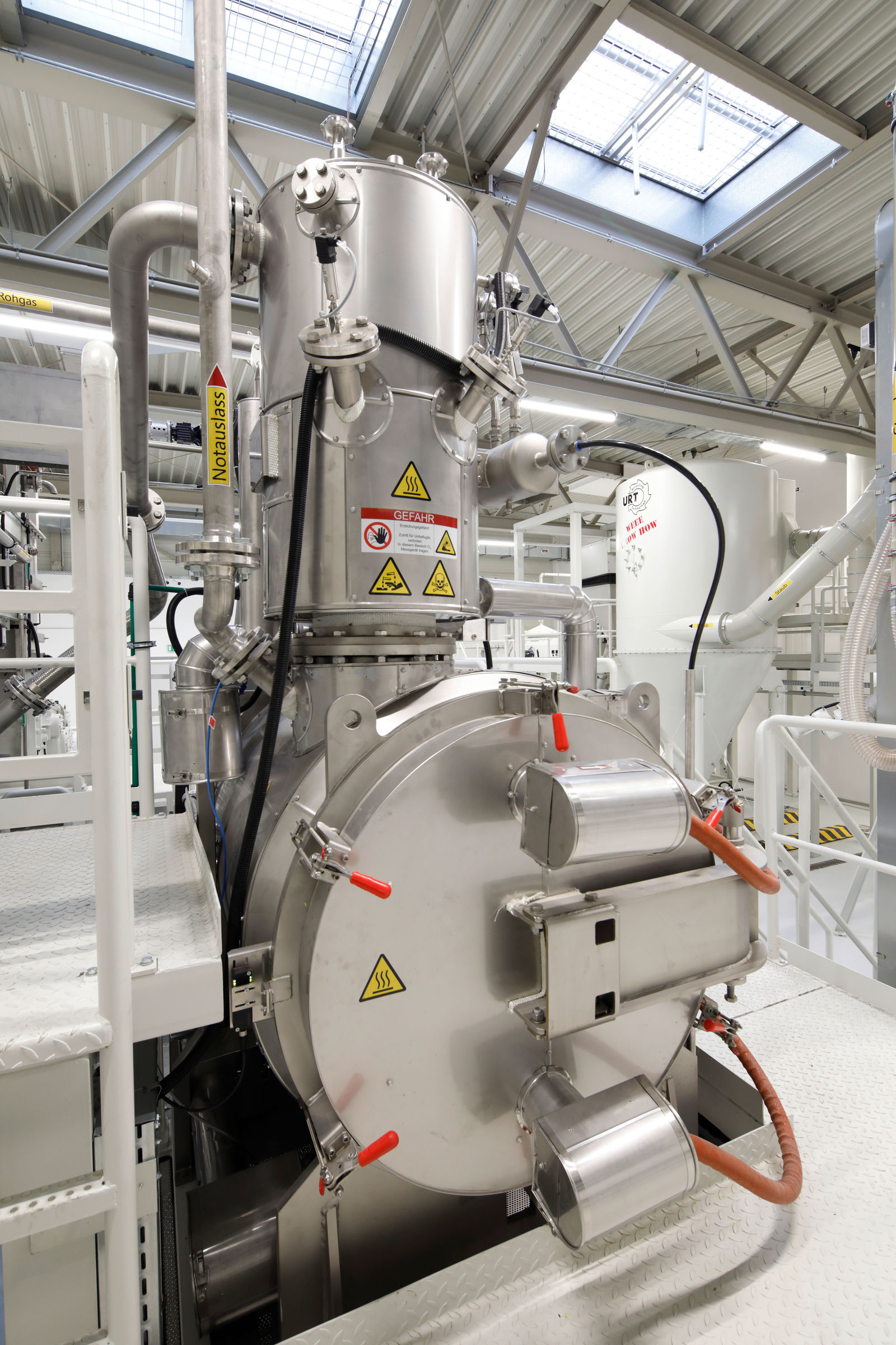 Industrielle Recyclinganlage mit einer großen, zylindrischen Metallkammer und mehreren angeschlossenen Rohren und Sicherheitsventilen. Warnschilder mit Gefahrenhinweisen wie „Heiß“ und „Gefahr“ sind angebracht. Im Hintergrund sind weitere Maschinen und eine helle Hallendecke mit Dachfenstern sichtbar.