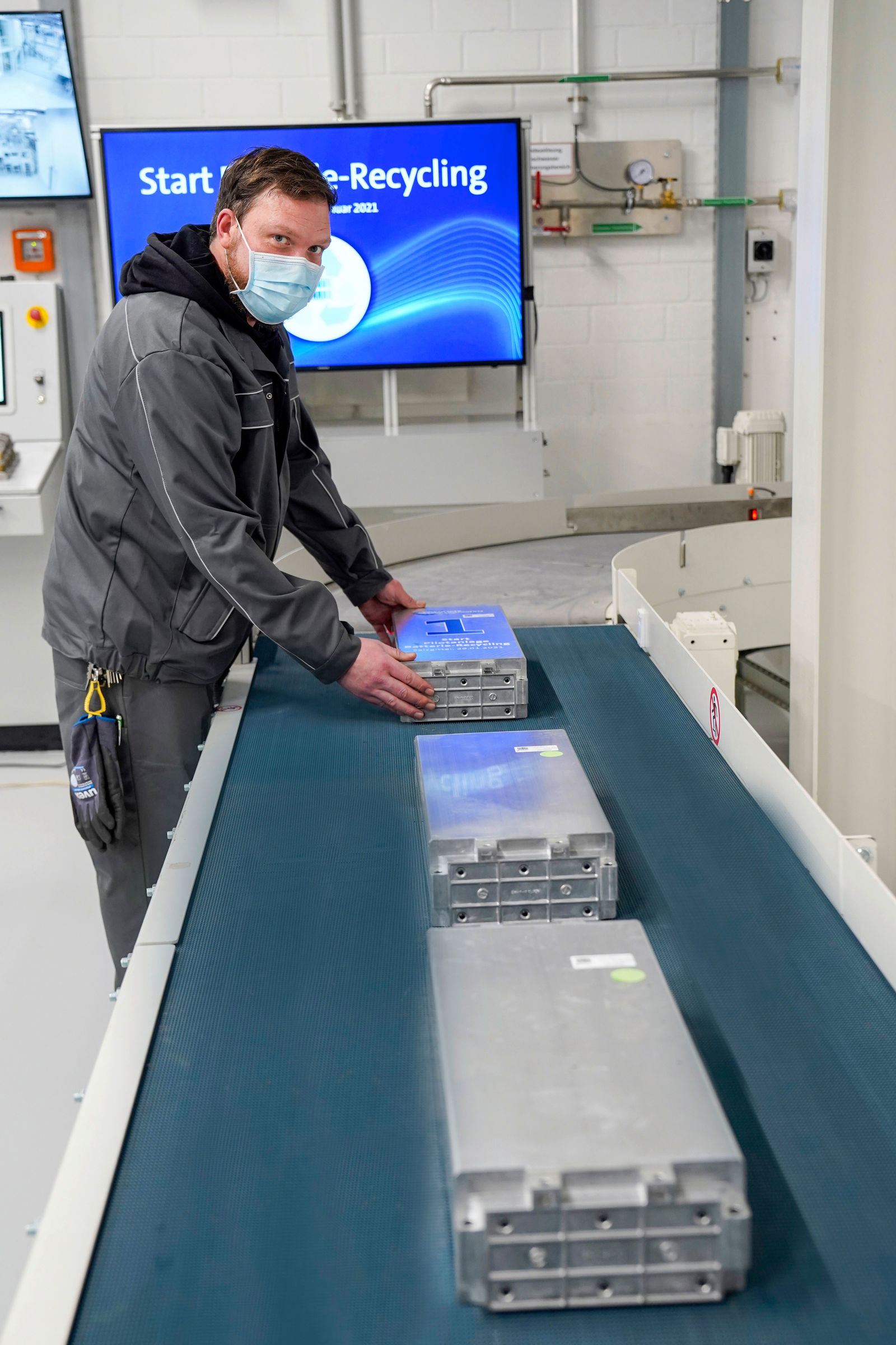 Ein Mitarbeiter mit Maske und grauer Arbeitskleidung platziert Batteriemodule auf einem Förderband in einer Recyclinganlage. Im Hintergrund zeigt ein Bildschirm die Aufschrift „Start Batterie-Recycling Januar 2021“.