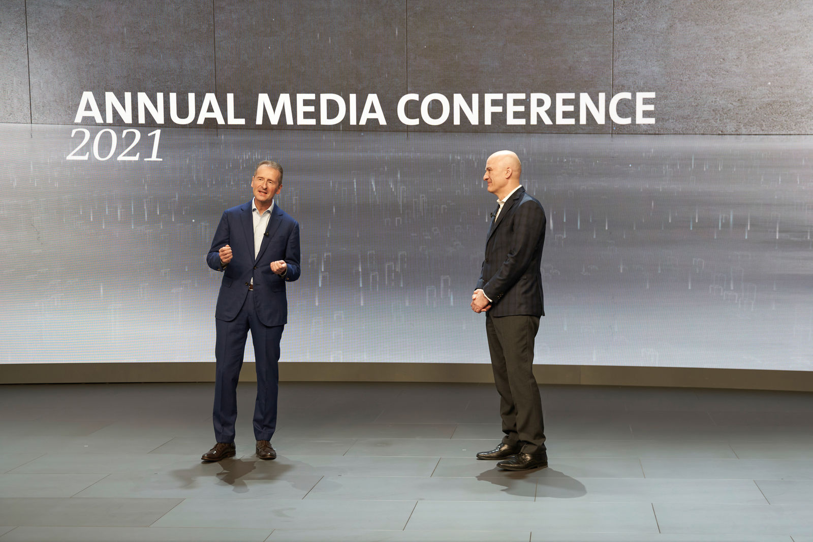 Two men stand on a stage and speak during the "Annual Media Conference 2021." The man on the left wears a dark blue suit and gestures while speaking. The man on the right wears a dark suit and listens attentively. A large screen in the background displays the conference title.