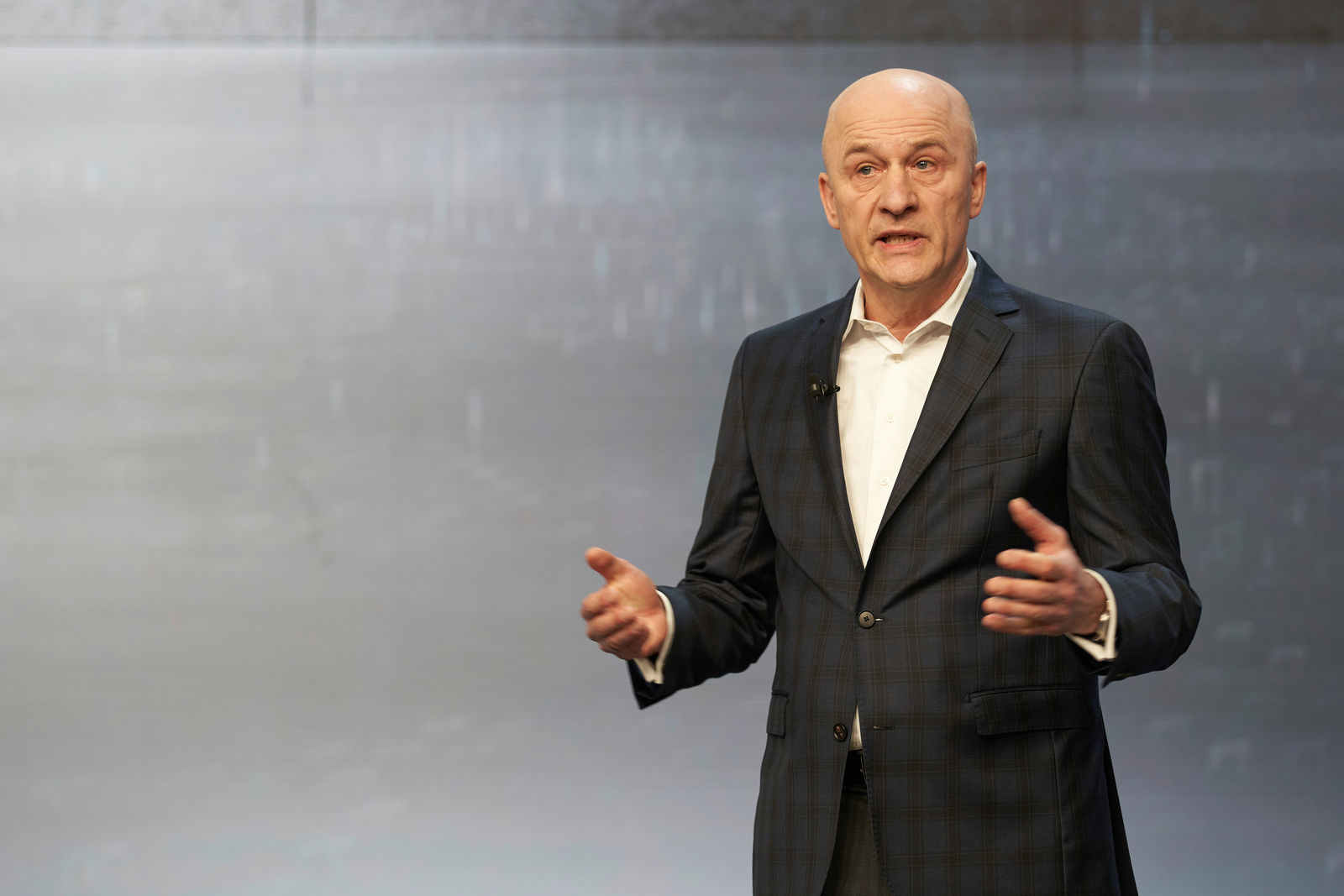 A man in a dark checkered suit speaks while gesturing on a stage. He is wearing a white shirt and has a microphone attached to his lapel. The background is neutral and modern.