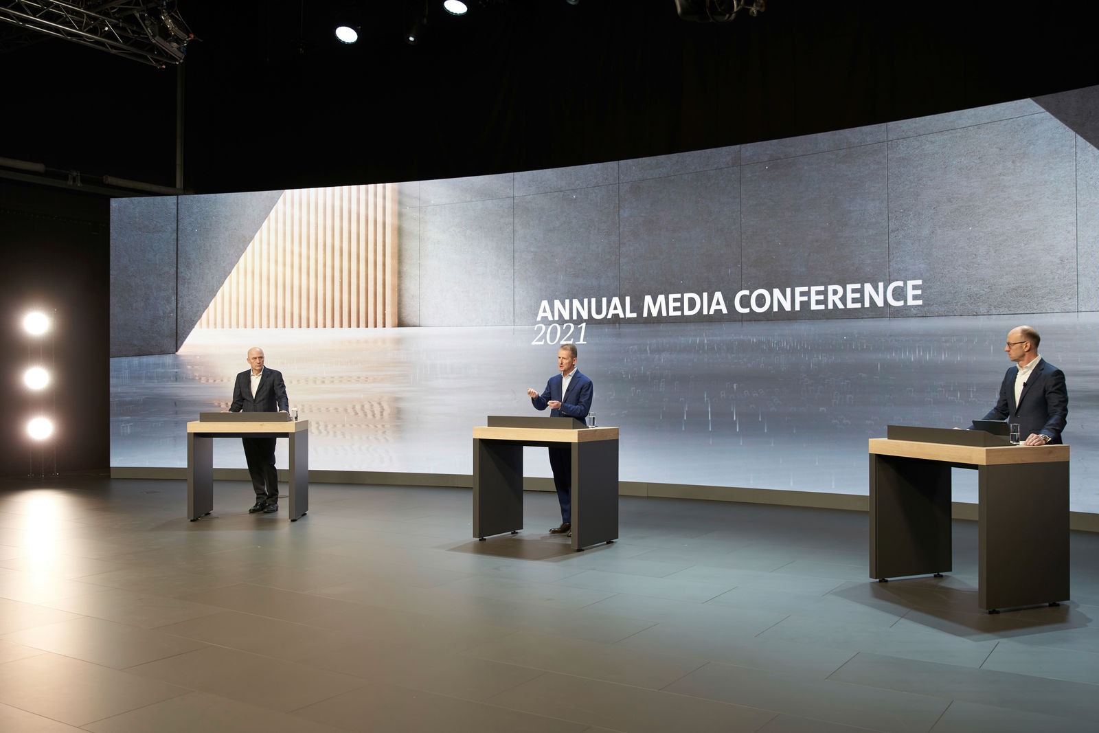 Three men stand at individual podiums on a stage during the "Annual Media Conference 2021." The man in the center wears a dark blue suit and gestures while speaking, while the other two men in dark suits listen attentively. A large screen in the background displays the conference title along with a modern graphic.