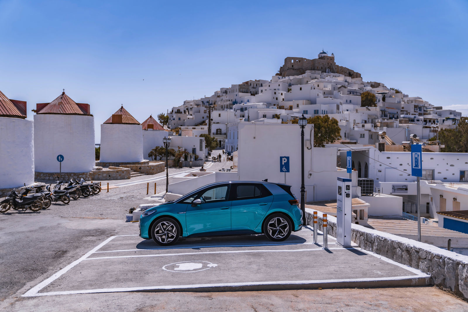 Ein blauer Volkswagen ID.3 steht auf einem Parkplatz mit Ladestation auf der griechischen Insel Astypalea. Im Hintergrund sind weiße Häuser, eine Festung auf einem Hügel und mehrere Windmühlen mit roten Dächern zu sehen. Der Himmel ist klar und blau.