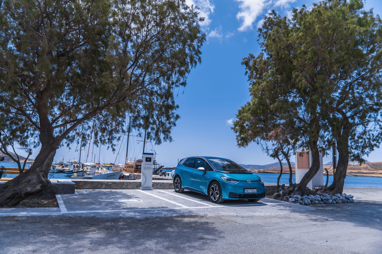 Ein blauer Volkswagen ID.3 steht an einer Ladestation auf einem Parkplatz am Meer, umgeben von Bäumen. Im Hintergrund liegen Segelboote im Hafen, und der Himmel ist strahlend blau mit wenigen Wolken.