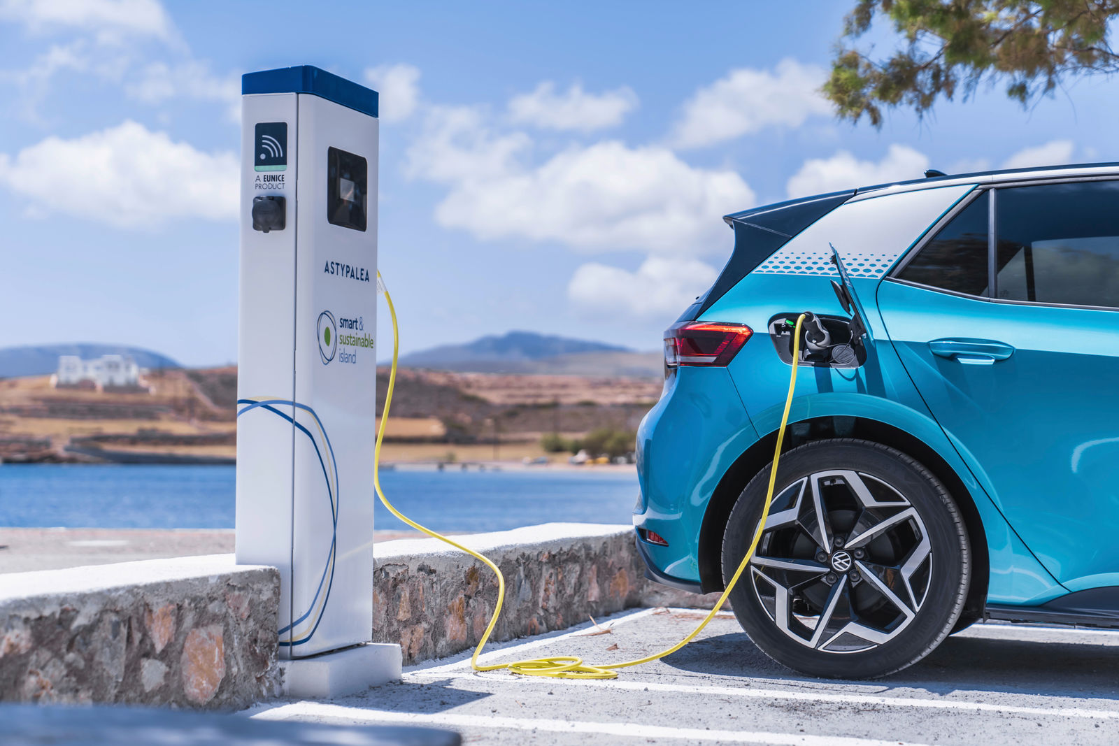 Ein blauer Volkswagen ID.3 lädt an einer E-Ladesäule auf der Insel Astypalea. Das gelbe Ladekabel ist mit der Ladestation verbunden. Im Hintergrund erstrecken sich eine hügelige Landschaft, das Meer und ein weißes Gebäude unter einem blauen Himmel mit wenigen Wolken.