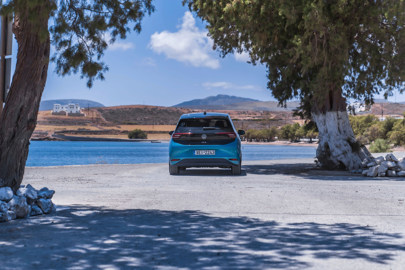 Ein blauer Volkswagen ID.3 steht mit Blick auf das Wasser auf einer sonnenbeschienenen Straße, die von zwei großen Bäumen eingerahmt wird. Im Hintergrund sind Hügel, ein weißes Gebäude und eine ruhige Bucht unter einem blauen Himmel mit wenigen Wolken zu sehen.