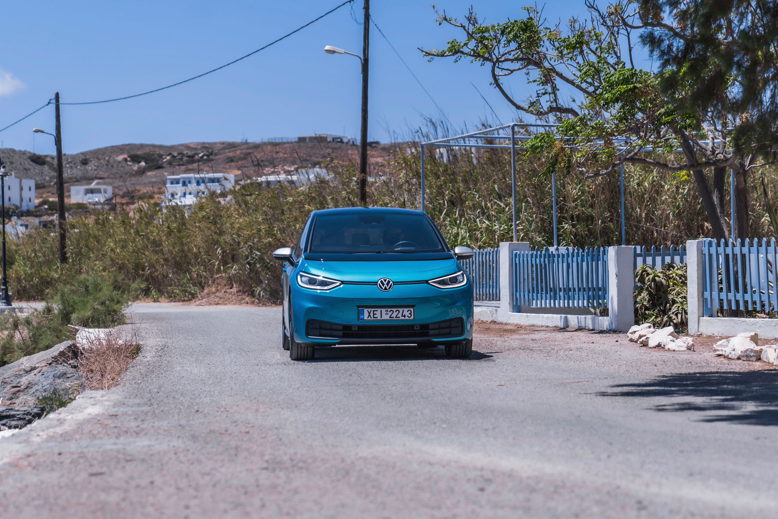 A blue Volkswagen ID.3 driving on a road alongside a white fence and surrounded by green bushes.