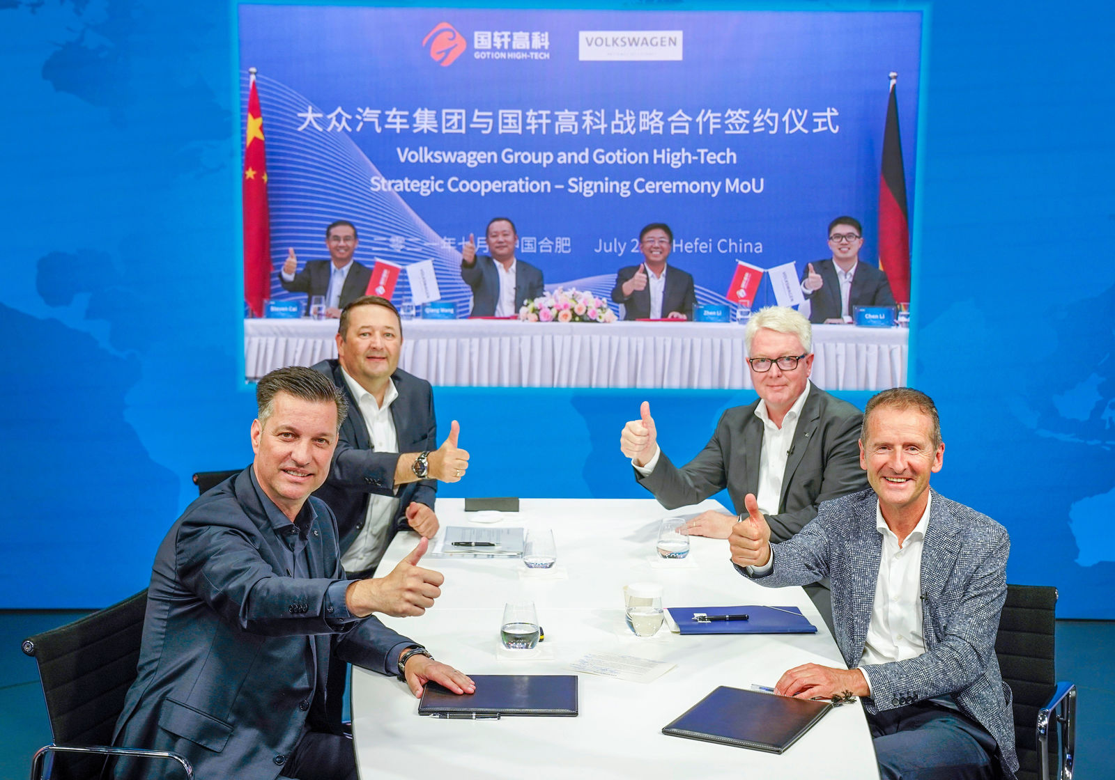 Four men are seated at a conference table, giving thumbs-up. In the background, a large screen displays the title "Volkswagen Group and Gotion High-Tech Strategic Cooperation – Signing Ceremony MoU." The screen also shows Chinese and German representatives participating in the virtual contract signing.