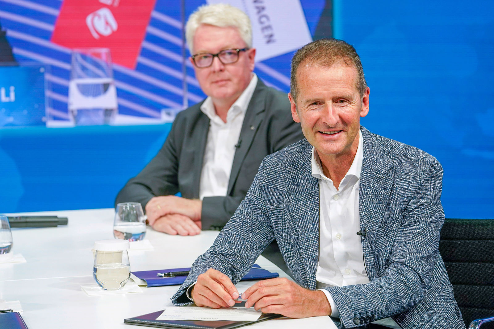 A man in a light blue blazer sits at a table, smiling at the camera. In front of him are a signed document folder and a glass of water. In the background, another man with white hair and glasses is also seated at the table, with a large digital display behind him featuring flags and logos.