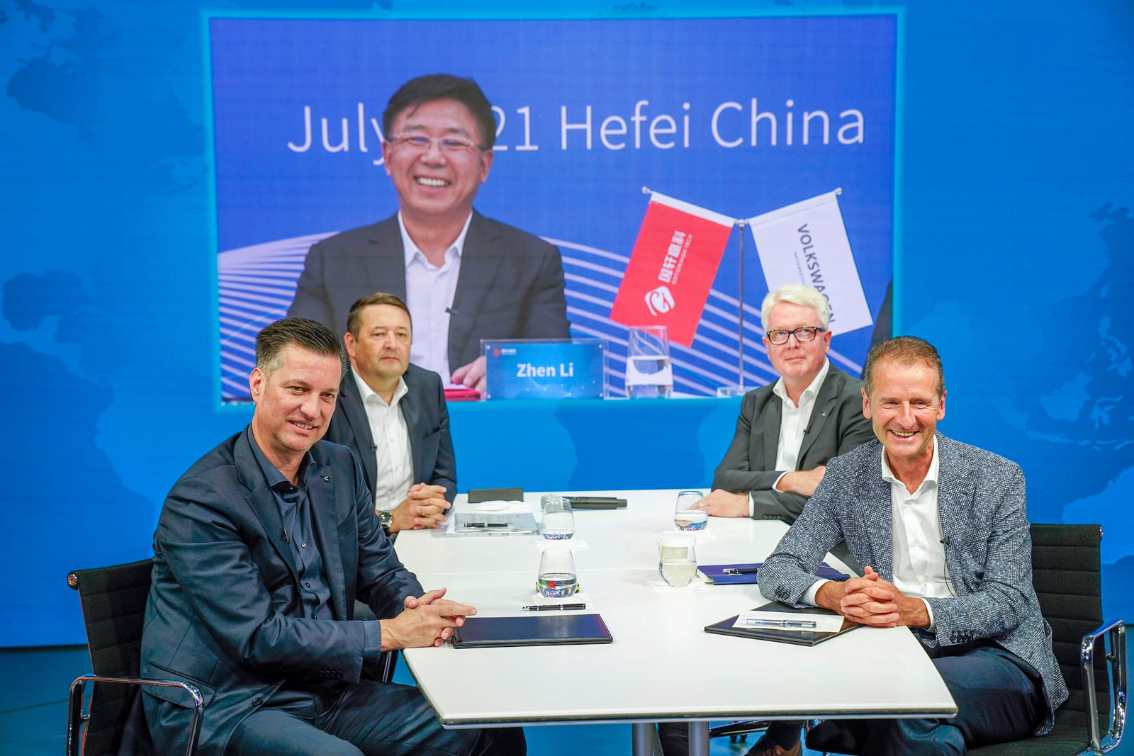 Four men are seated at a modern conference table with notebooks, pens, and glasses of water. They are dressed in business attire and smiling at the camera. In the background, a large screen displays a virtual participant from China, who is also smiling. The screen features the words "July 21 Hefei China" and the name "Zhen Li". Two small flags with the logos of Volkswagen and a Chinese company are placed in front of him. The setting appears to be a professional studio or conference room with a world map as a backdrop.