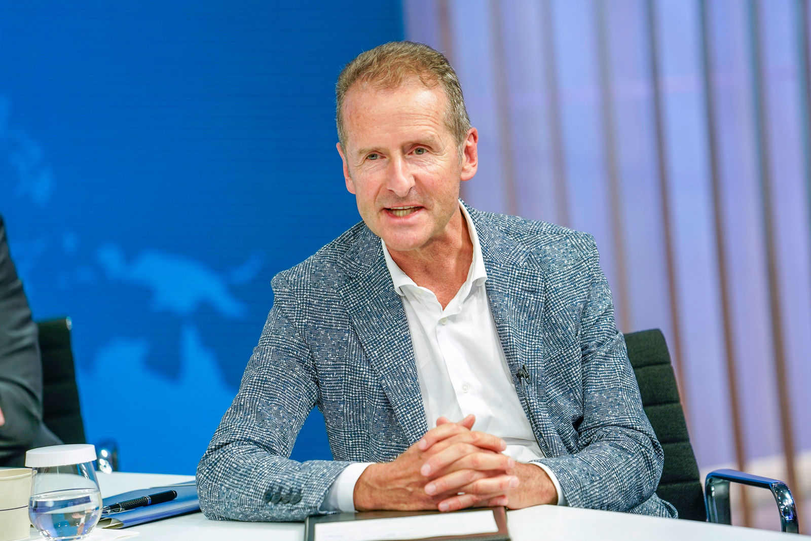 A middle-aged man with short, slightly graying hair sits at a table in a modern conference room or television studio. He wears a white shirt and a textured blazer. His hands are clasped, and he is either speaking or attentively listening. In the background, a blue world map on a screen and semi-transparent curtains are visible.