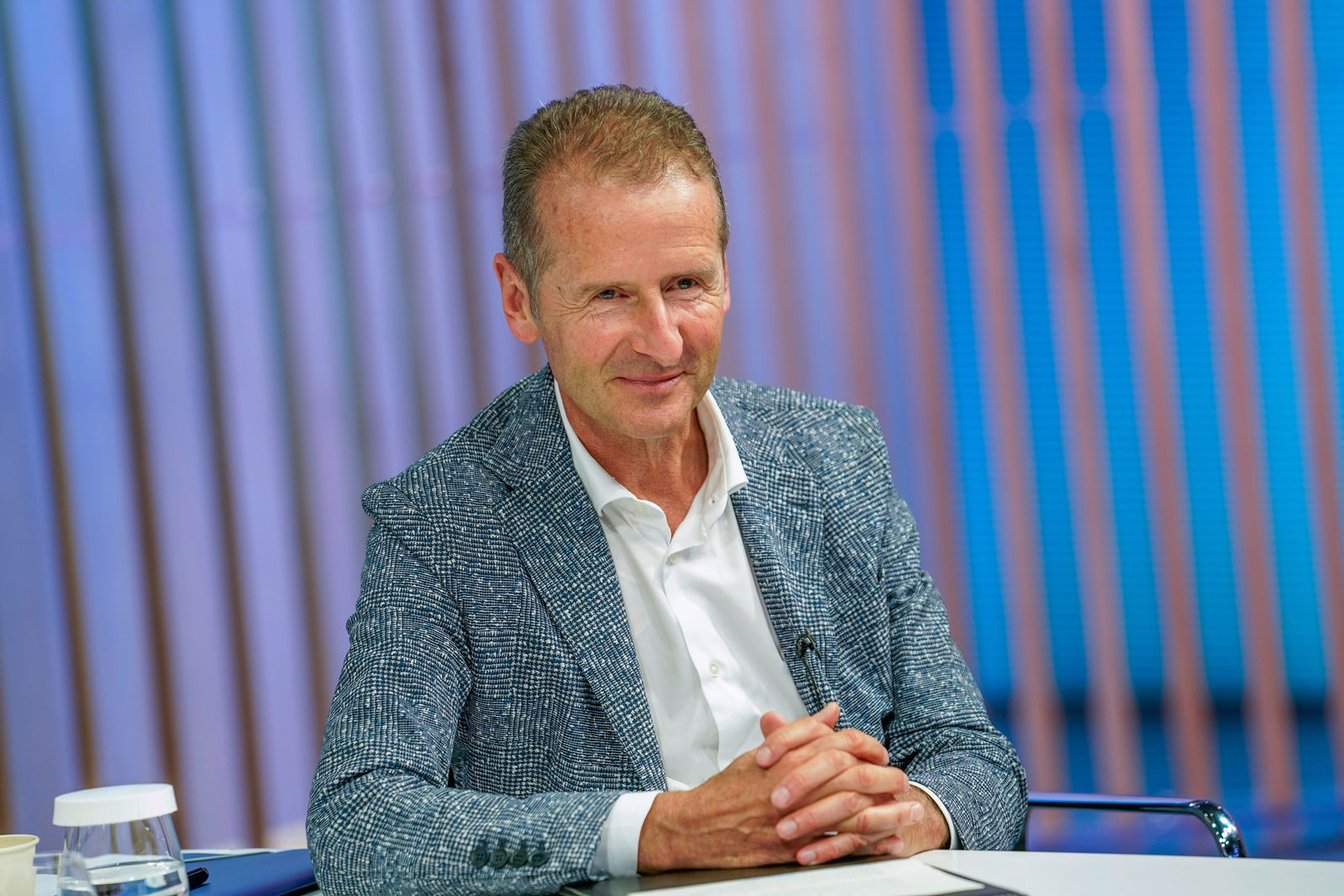 A middle-aged man with short, slightly graying hair sits at a table, smiling warmly. He is wearing a white shirt under a patterned blue blazer. The background features vertical blue and orange light strips. On the table in front of him, there is a glass of water and some documents.