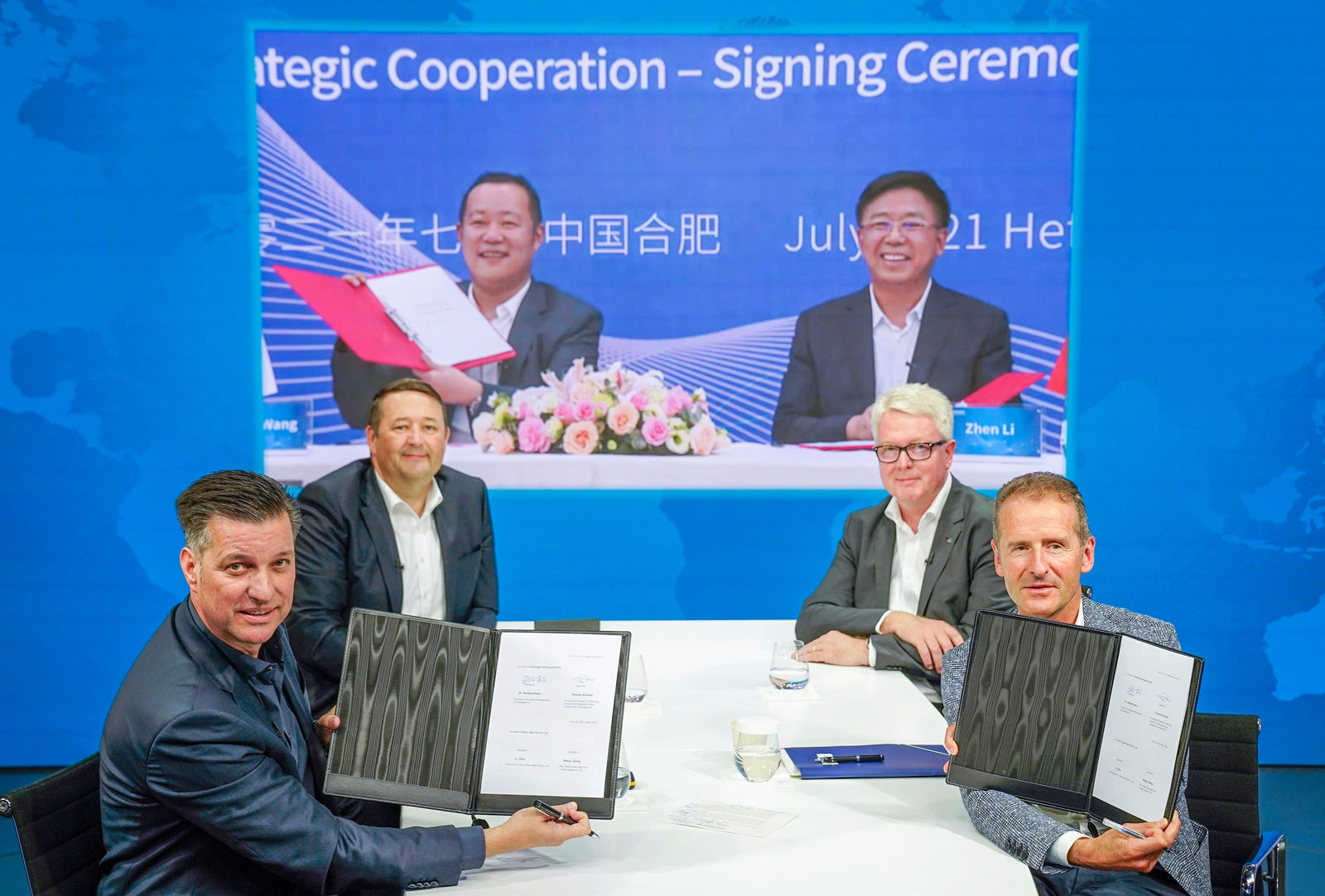 Four men are seated at a table, with two of them holding open document folders displaying signed agreements. In the background, a large screen shows two more men also holding signed documents. The banner on the screen announces a strategic cooperation between Volkswagen Group and Gotion High-Tech.