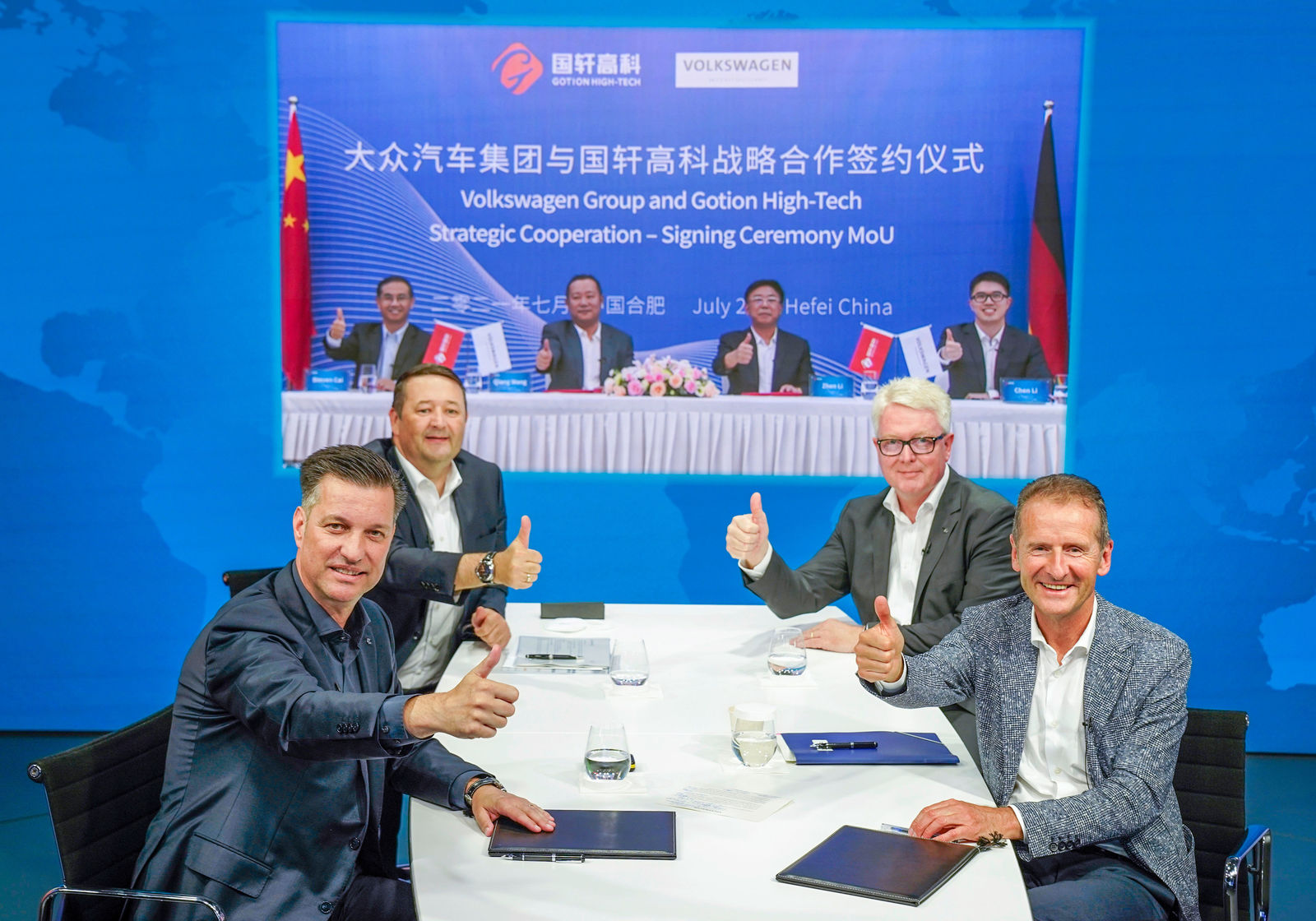 Four men are seated at a table, giving a thumbs-up gesture. In front of them are document folders and glasses of water. In the background, a large screen displays a blue banner announcing the signing of a Memorandum of Understanding (MoU) between Volkswagen Group and Gotion High-Tech. The banner also shows four additional men giving a thumbs-up.