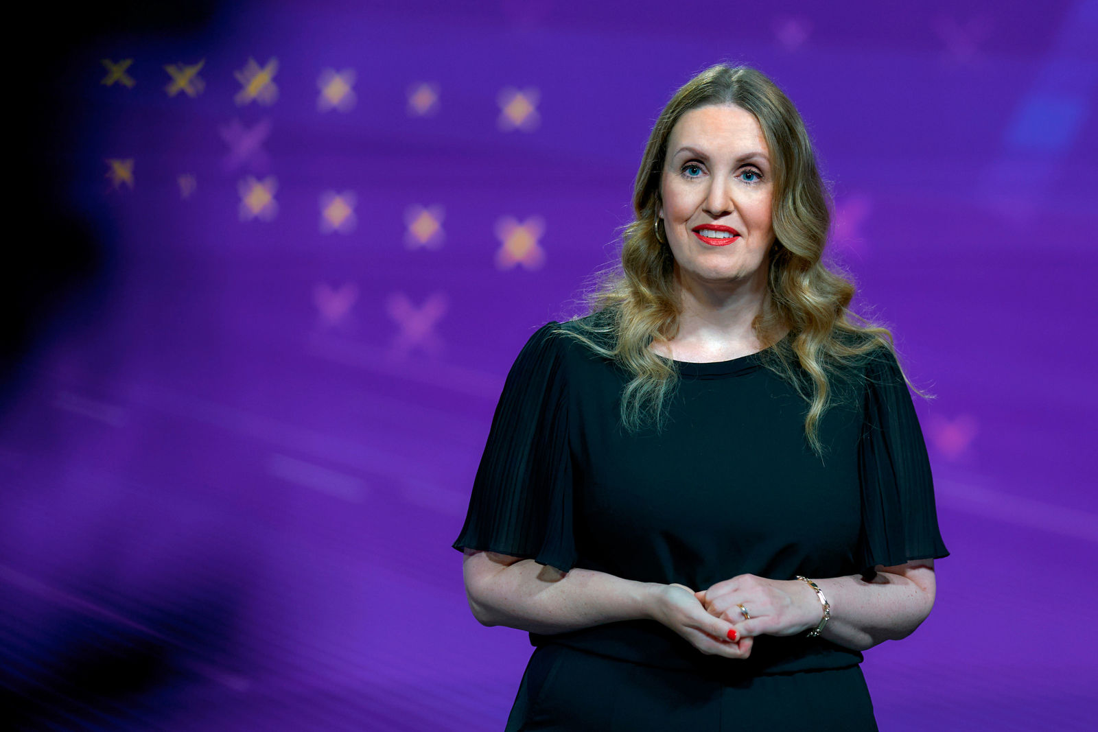 Eine Frau mit langen, welligen blonden Haaren steht vor einem violetten Hintergrund mit stilisierten gelben und rosa Kreuzen. Sie trägt ein schwarzes Kleid mit kurzen, weiten Ärmeln und hält ihre Hände locker vor sich zusammen. Sie lächelt leicht und blickt in Richtung des Publikums.
