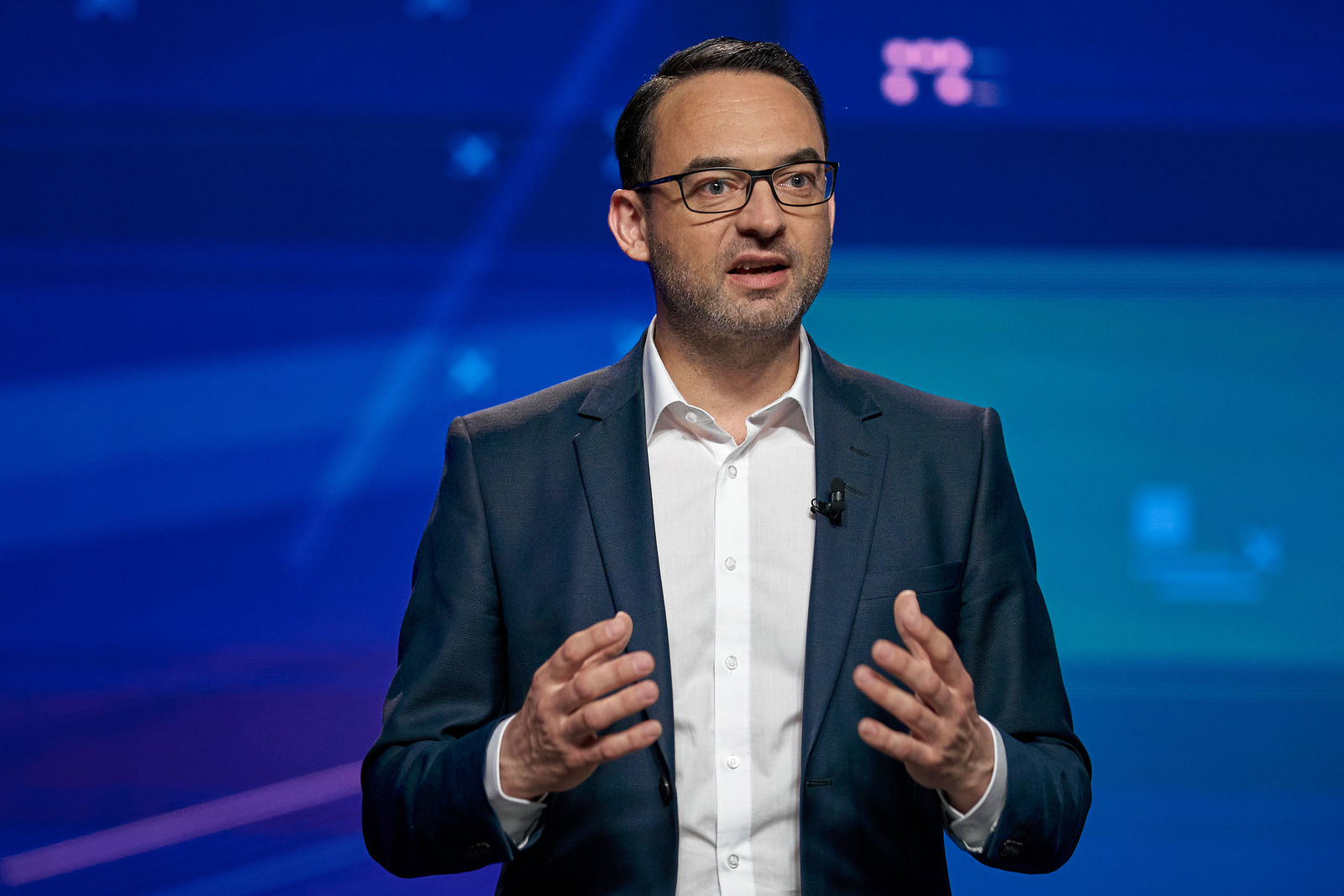 Ein Mann mit dunklem Haar, Brille und einem dunkelblauen Anzug spricht auf einer Bühne. Er trägt ein weißes Hemd ohne Krawatte und hat ein Mikrofon am Revers befestigt. Der Hintergrund ist eine digitale blaue Leinwand mit grafischen Elementen.