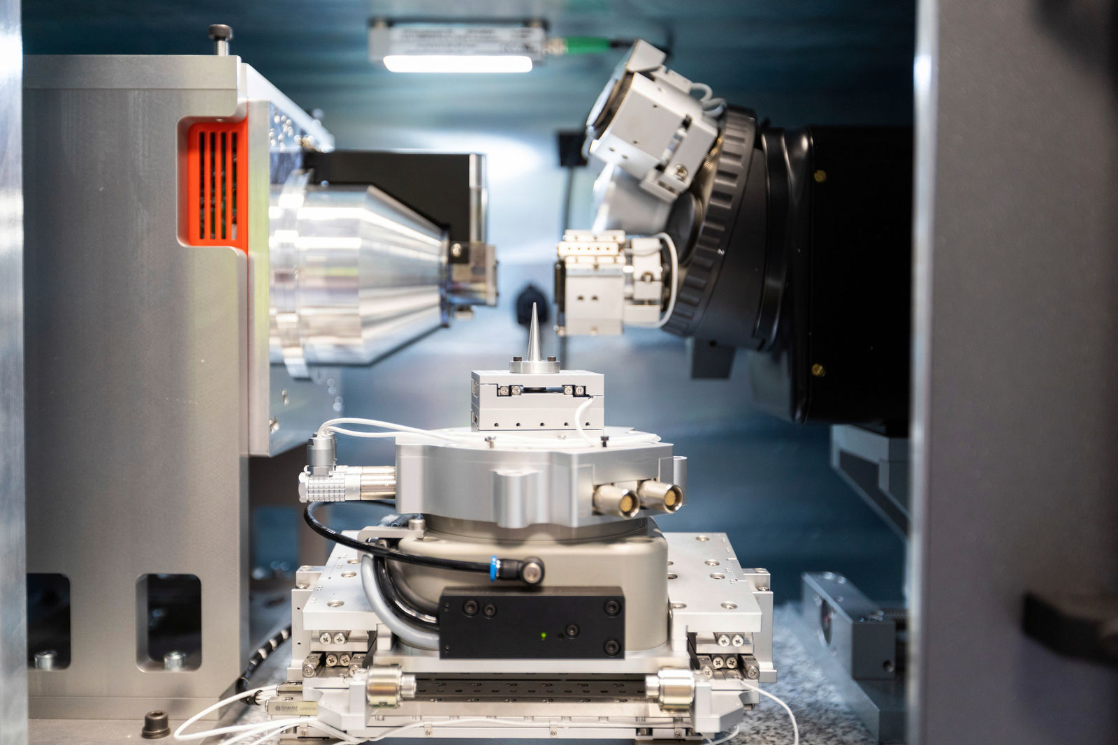 Close-up of a high-precision scientific or industrial measurement device with multiple optical and mechanical components. The equipment features metal housings, cable connections, and movable parts for precise analysis or testing.