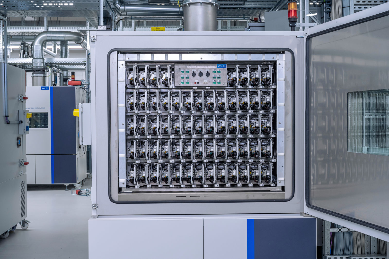 An open high-tech test chamber with numerous battery test modules in a modern laboratory setting. The device has a control panel with indicators and buttons. Additional technical equipment is visible in the background.