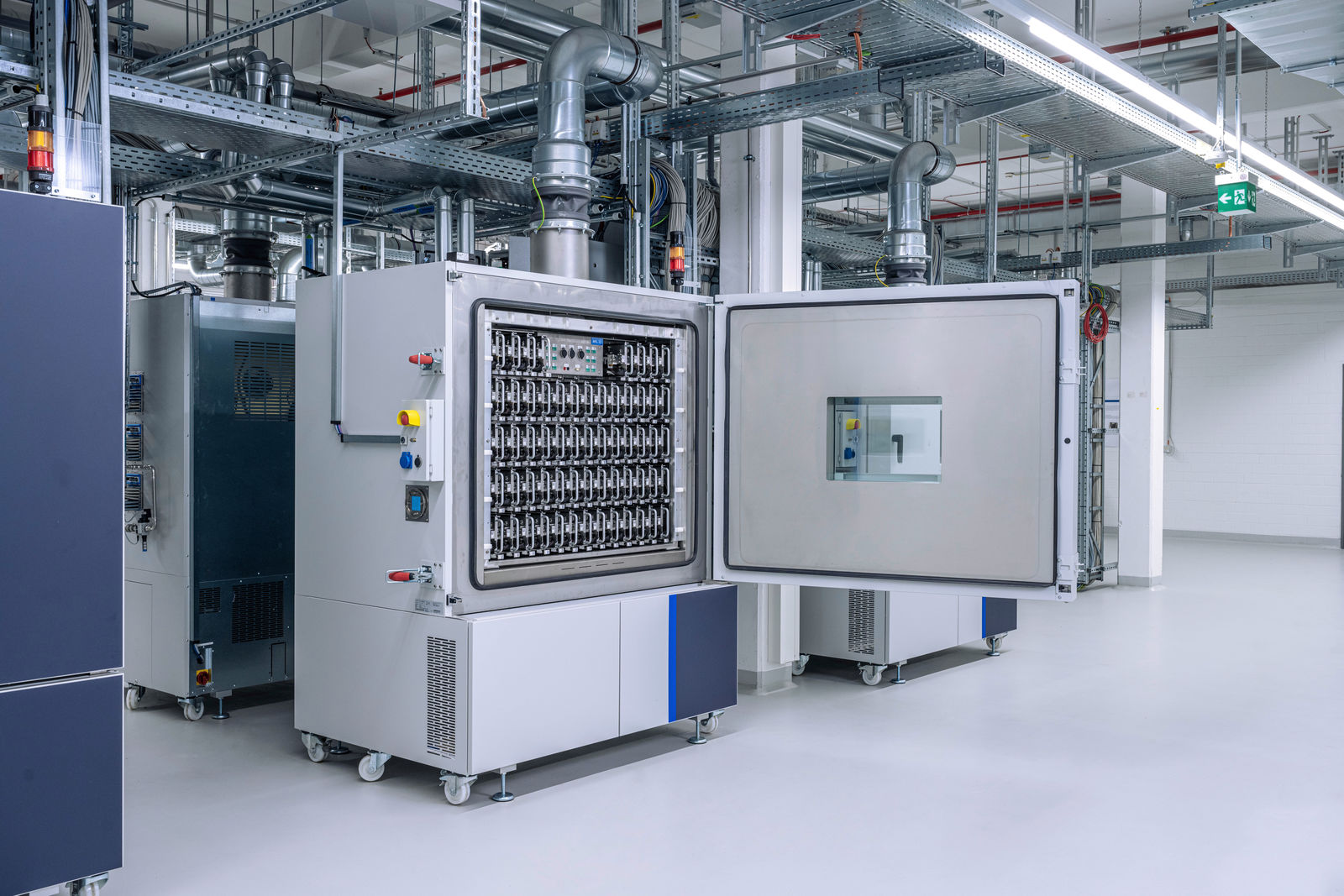 A high-tech laboratory featuring an open climate chamber with multiple battery cells neatly arranged inside. The chamber is connected to a complex ventilation and cooling system. The environment is clean, with white walls and industrial metal structures on the ceiling.
