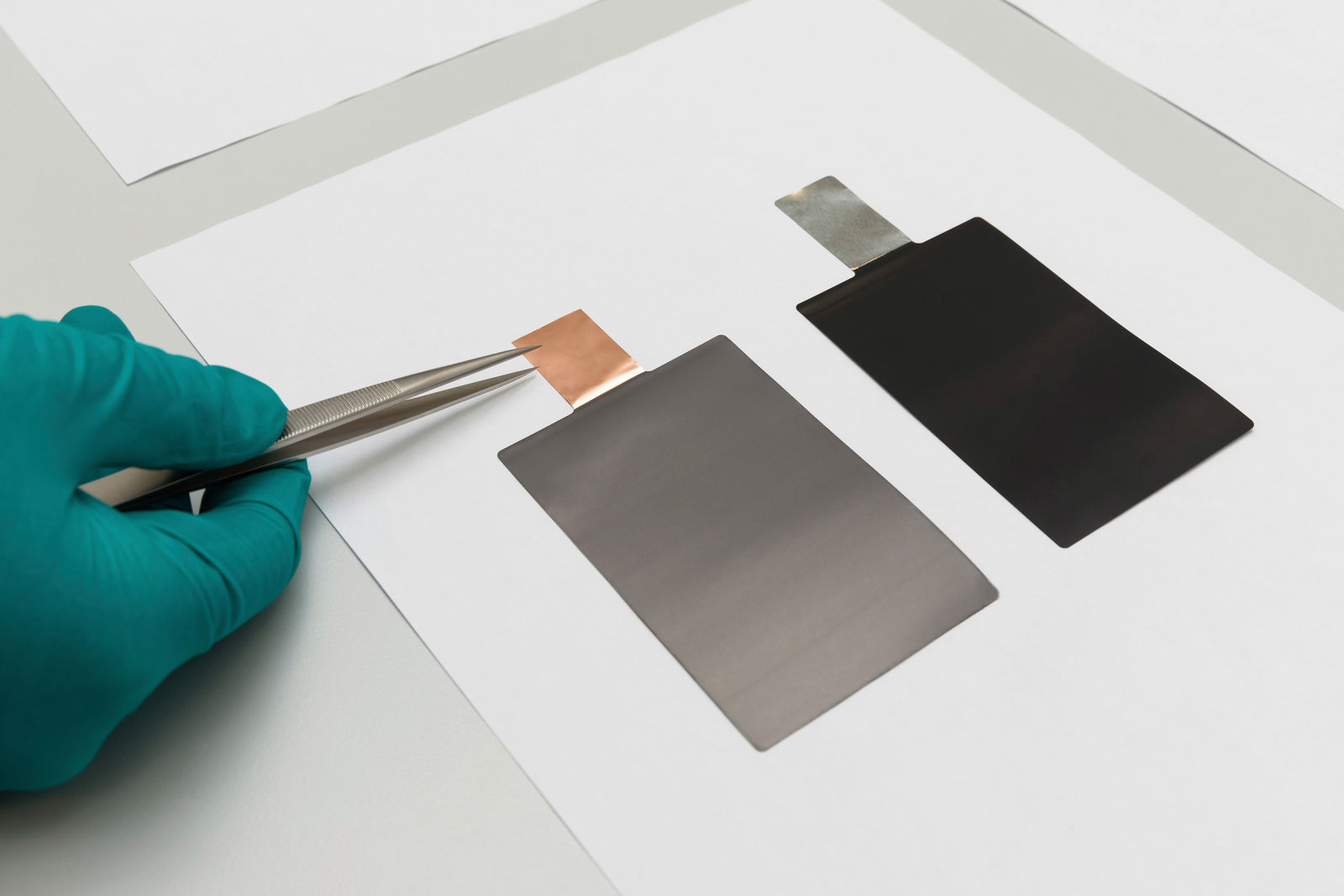 Close-up of a hand wearing blue protective gloves, using tweezers to hold a metallic battery cell. Two rectangular battery cells lie on a white sheet of paper – one with a copper-colored tab and the other with a silver-colored tab.