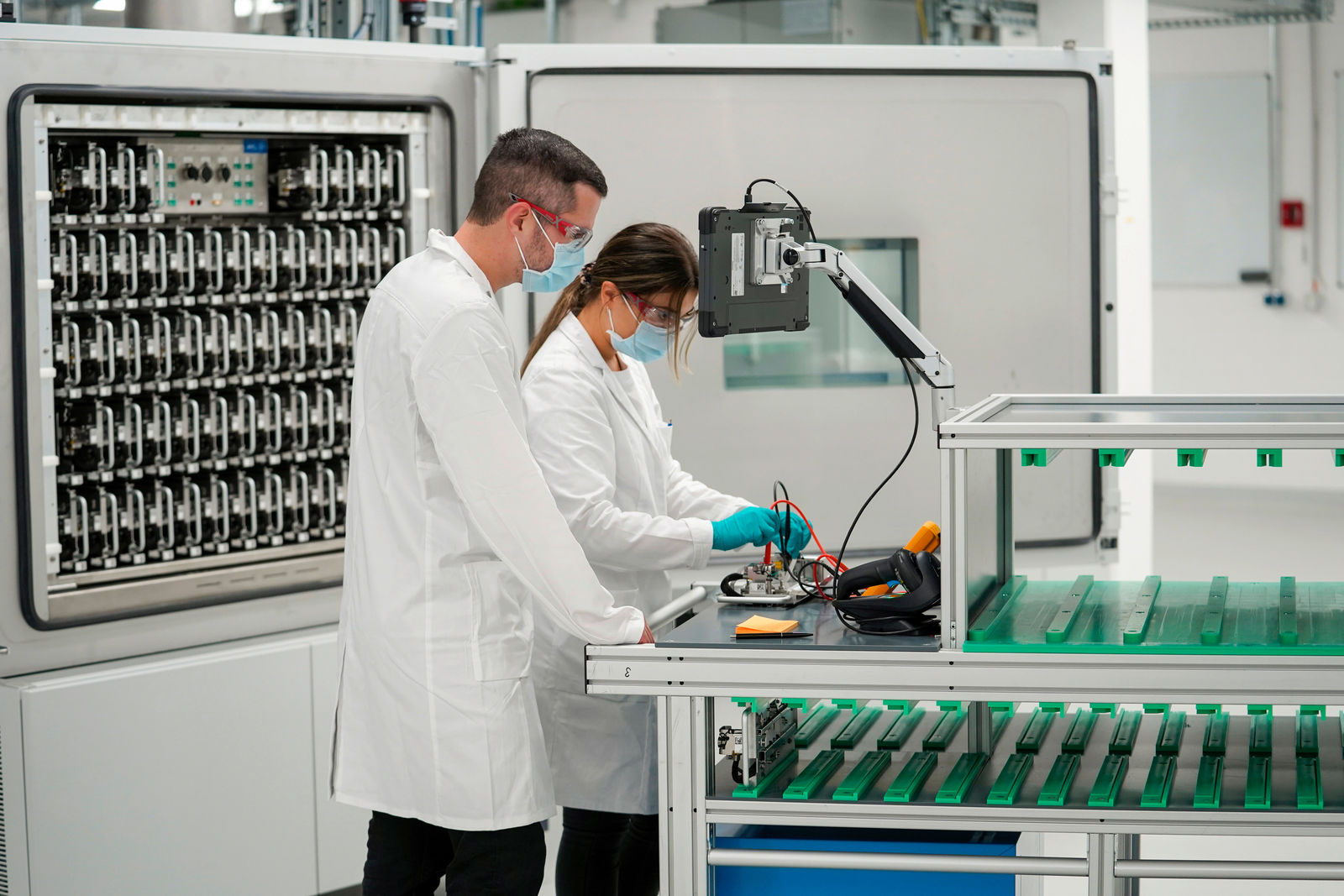 Two people in lab coats work in a lab on a battery, surrounded by machines and technical equipment.