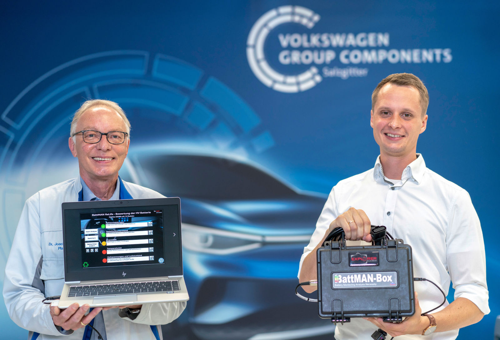 Two men stand in front of a Volkswagen Group Components backdrop. One holds a laptop, and the other a portable box.
