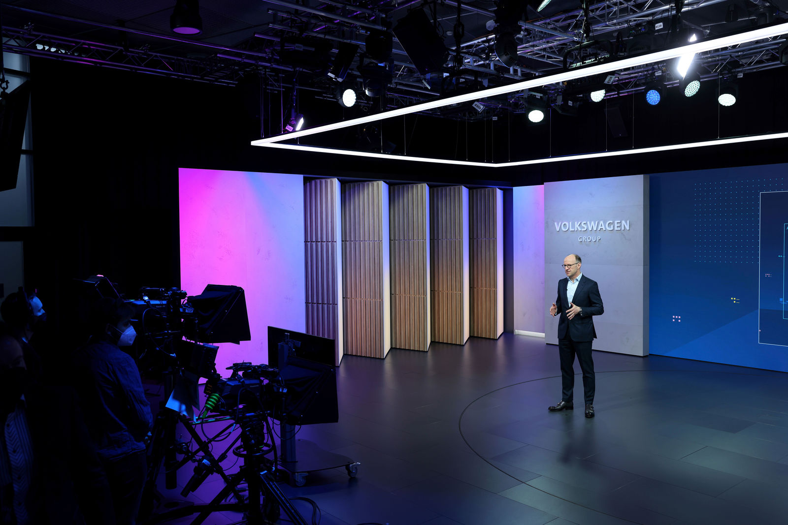 A man in a suit and glasses stands on a stage with Volkswagen branding, delivering a presentation. The stage has a modern design with blue and purple lighting accents and a textured backdrop. In the foreground, cameras and a production team are recording the event.