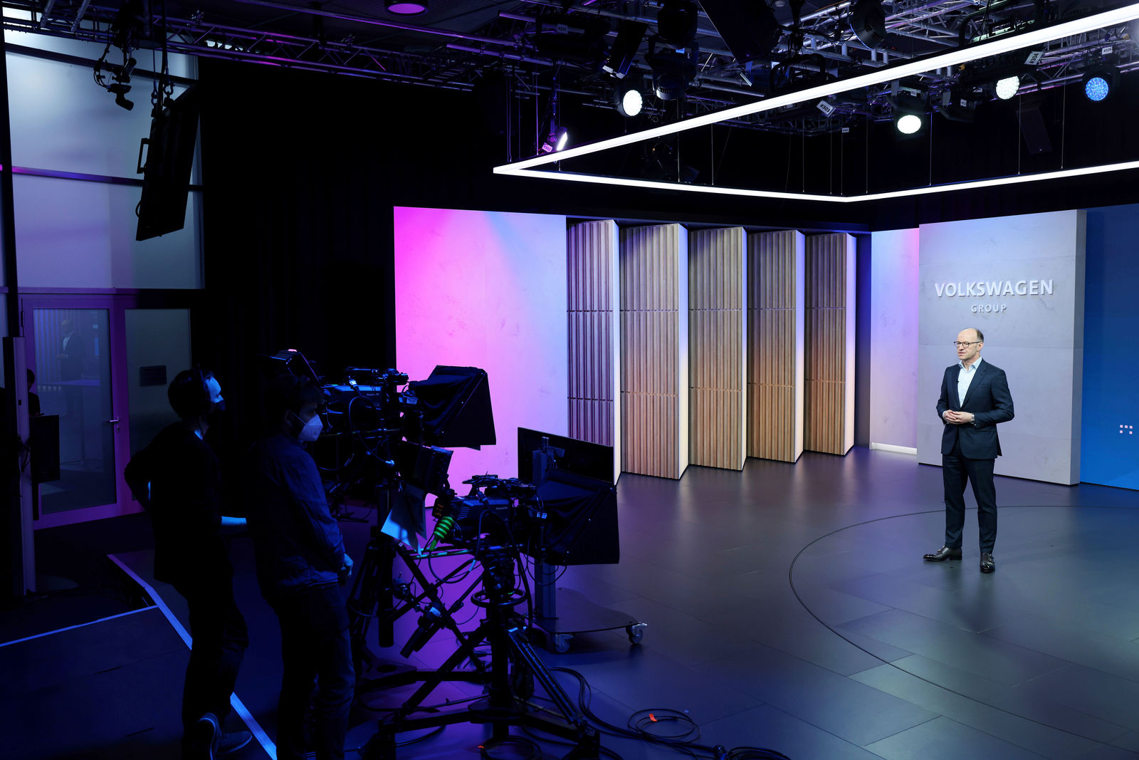 A man in a dark suit speaks on a modern stage with the "Volkswagen Group" logo. On the left, a camera crew films the scene with multiple cameras. The studio lighting features blue and purple accents.