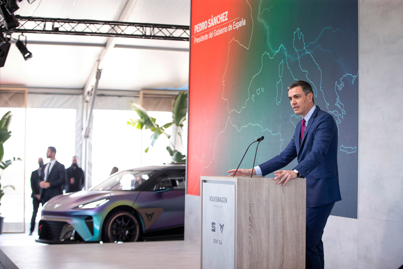 Spanish Prime Minister Pedro Sánchez delivers a speech at a podium featuring Volkswagen and SEAT logos. In the background, a large wall displays a map of Europe along with his name in red letters. Beside him, a futuristic car with a sleek design and iridescent paint is visible. Security personnel and other attendees can be seen in the background.