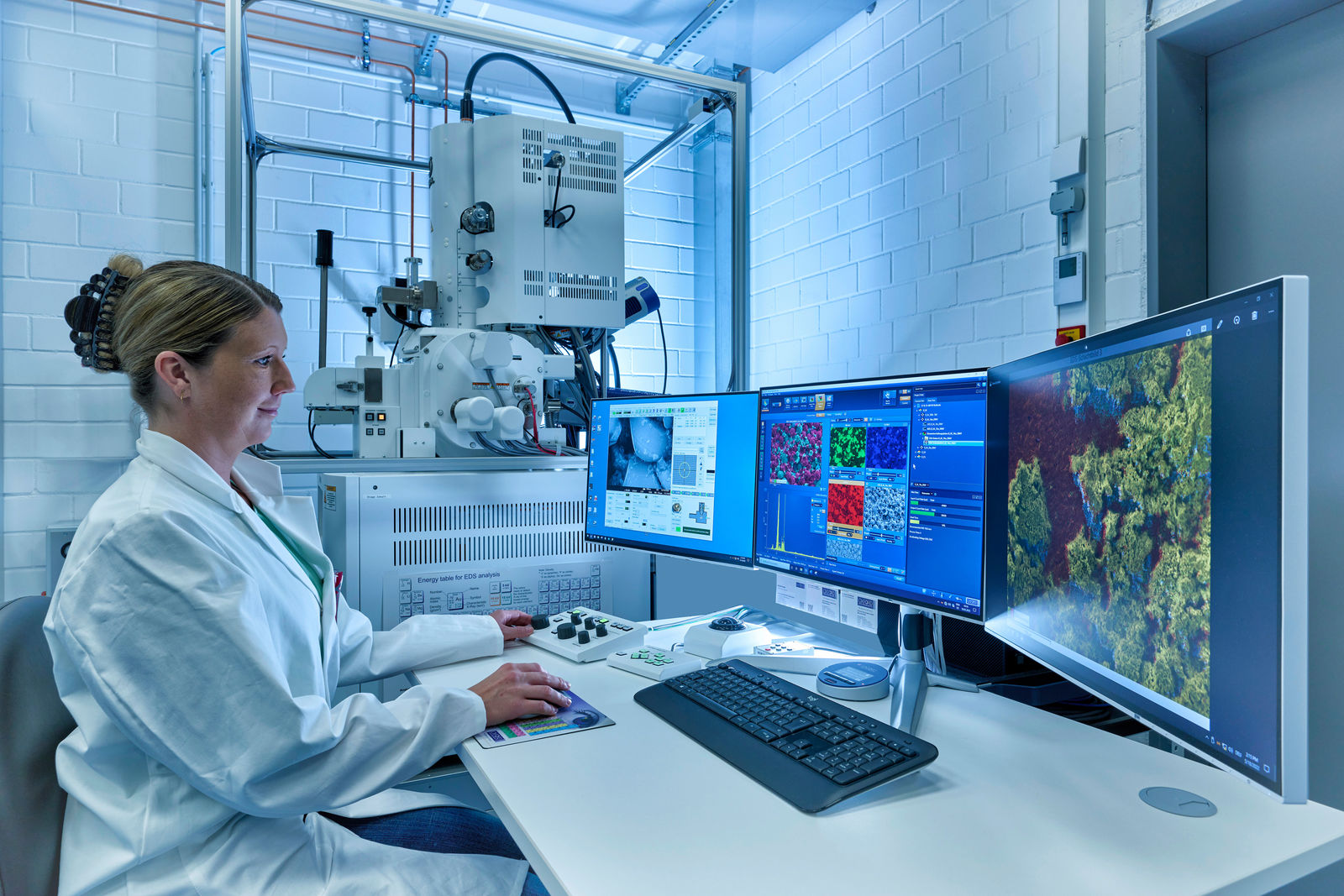 Eine Wissenschaftlerin in einem weißen Laborkittel analysiert Mikroskopiebilder an mehreren Bildschirmen in einem High-Tech-Labor.