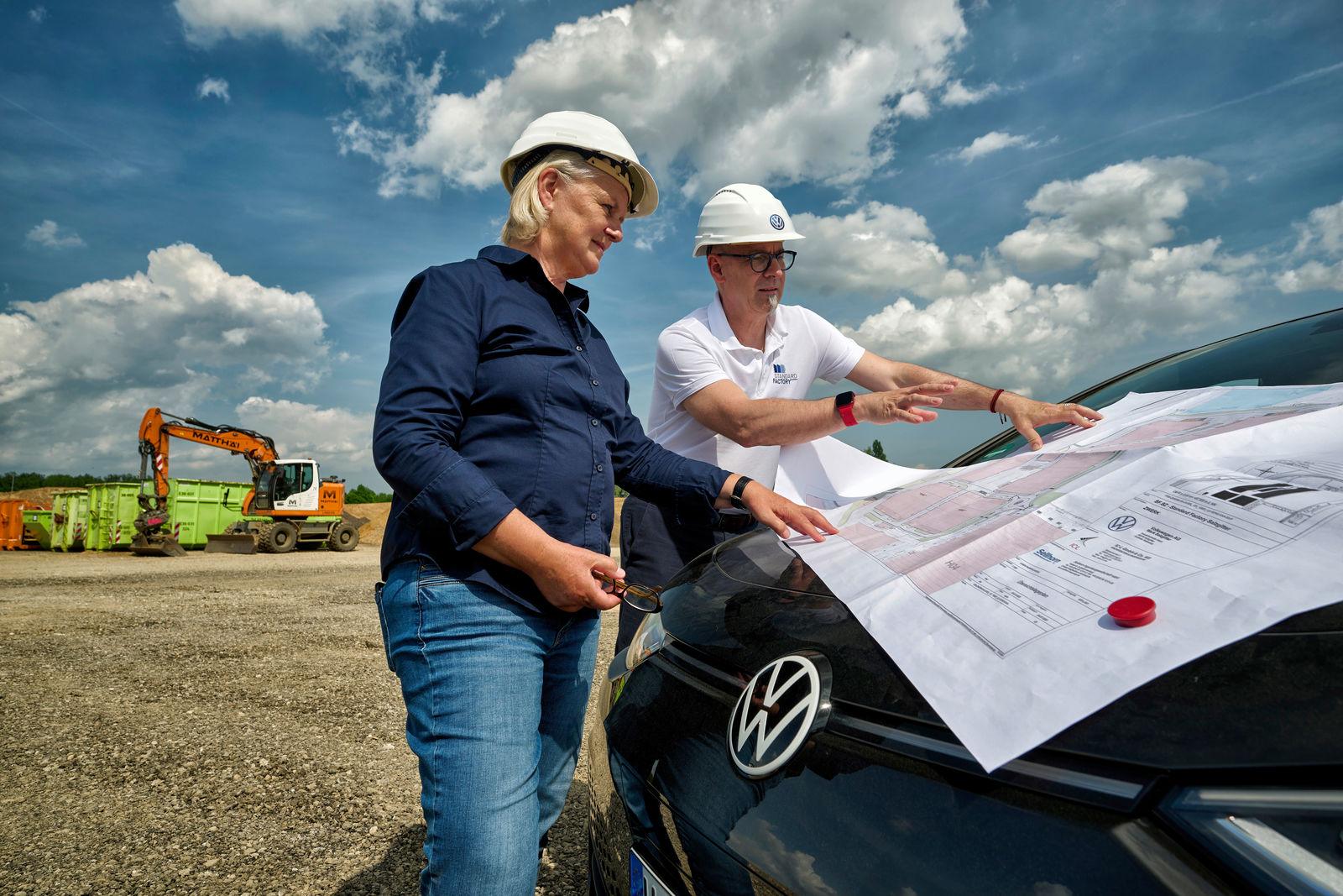 Zwei Bauleiter mit Schutzhelmen besprechen Baupläne, die auf der Motorhaube eines Volkswagen-Autos ausgebreitet sind.
