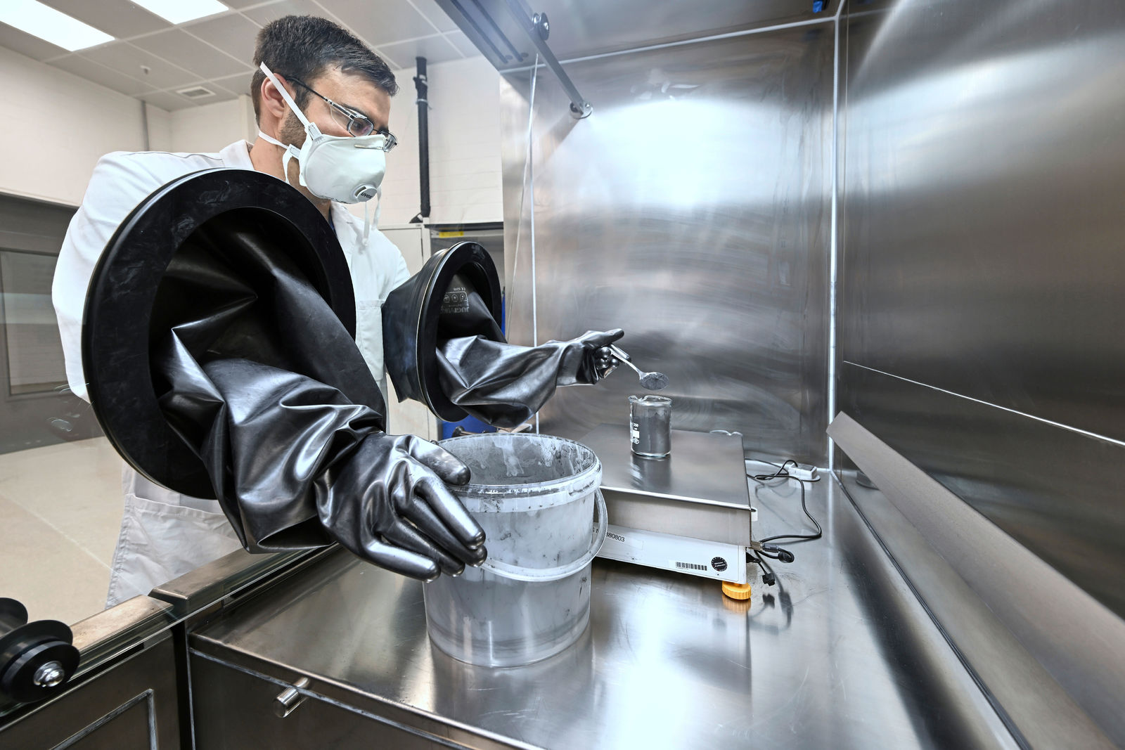 A scientist in protective clothing, including a white lab coat, face mask, and safety glasses, is working inside a stainless-steel glove box. He is using fixed black rubber gloves to handle a substance in a transparent container. The sterile environment features a precision scale and reflective metal walls.
