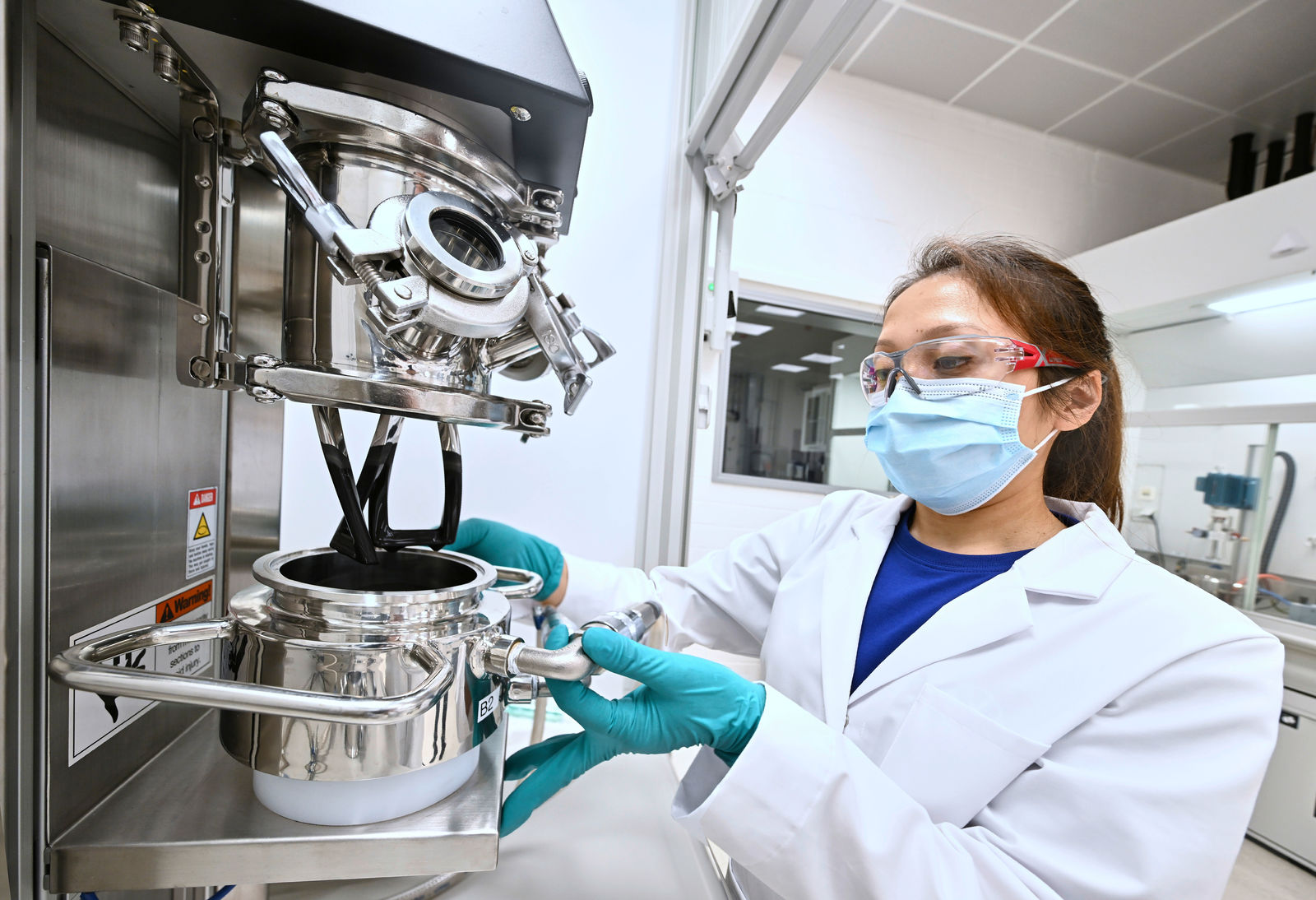 Eine Wissenschaftlerin in einem weißen Laborkittel, Schutzbrille, Mundschutz und blauen Schutzhandschuhen arbeitet in einem modernen Labor. Sie bedient eine große, hochglanzpolierte Metallmaschine, aus der eine schwarze, viskose Substanz in einen darunterliegenden Behälter fließt. Im Hintergrund sind weitere Laborgeräte und eine Glasscheibe zu sehen.