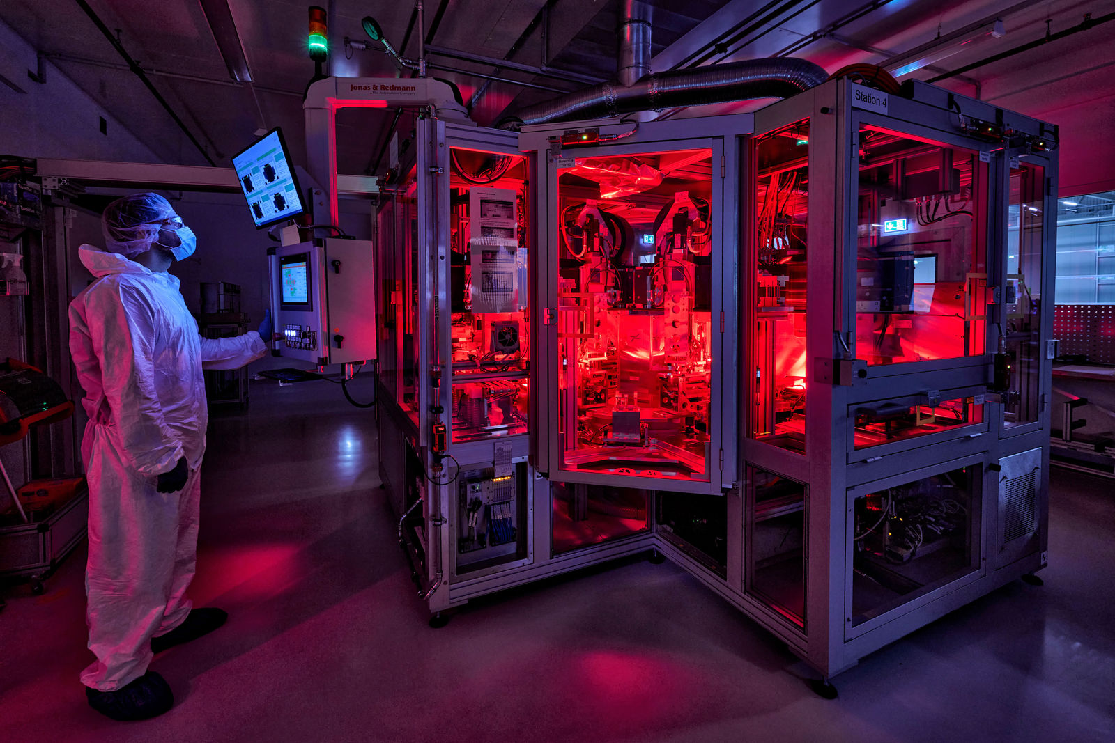 Ein Mitarbeiter in Schutzkleidung überwacht eine rot beleuchtete Hightech-Maschine in einer Reinraumumgebung.