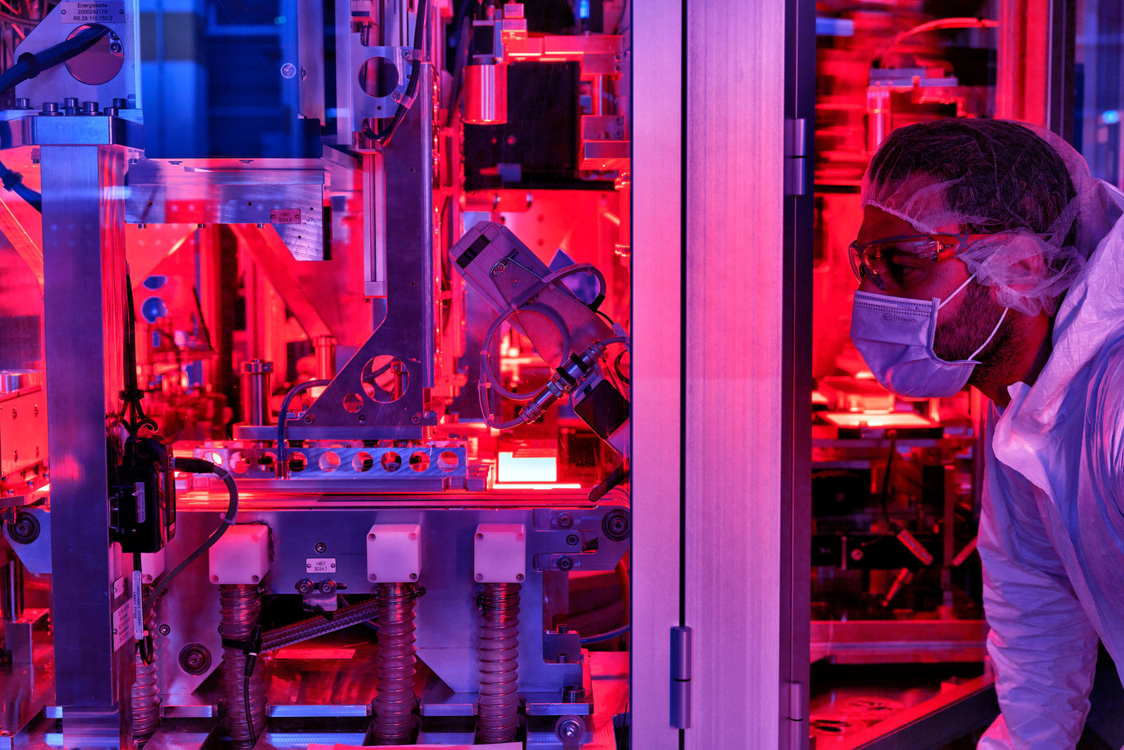 The image shows a technician working in a state-of-the-art manufacturing facility. The red lighting and precise equipment suggest advanced manufacturing technology, possibly in the field of battery cell production or electronics manufacturing.