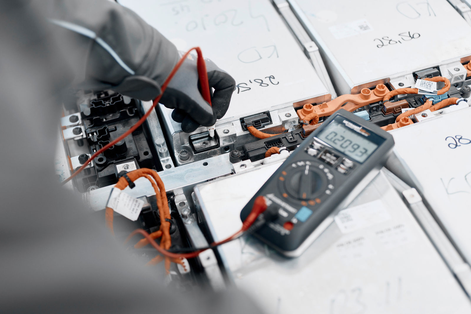 Nahaufnahme einer Person in Schutzkleidung, die mit einem Multimeter die Spannung einer Batteriezelle misst. Die Batterieeinheiten sind mit orangen Hochvolt-Kabeln verbunden und mit handschriftlichen Notizen markiert.