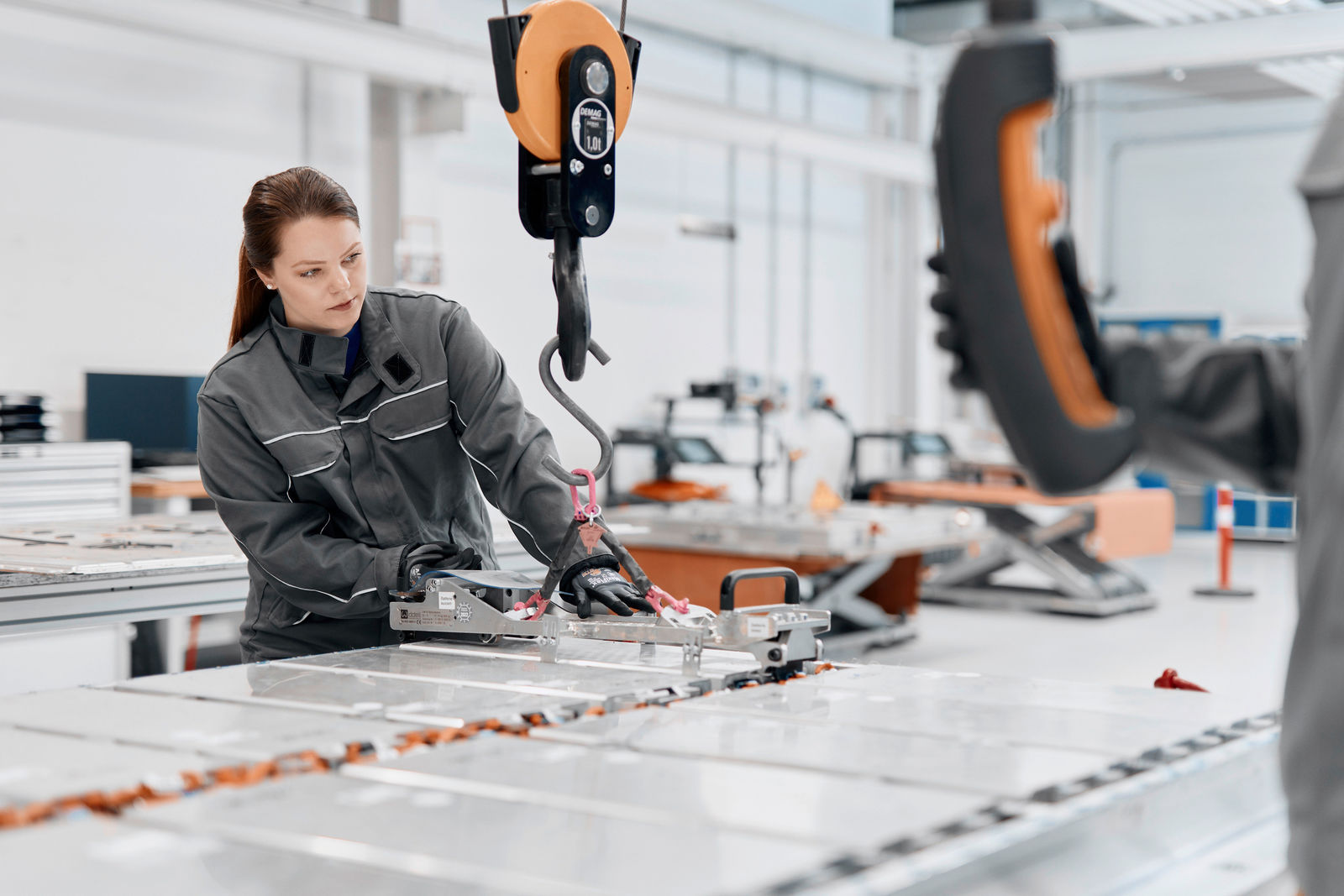 A worker in gray protective clothing assembles a large battery cell using a hoist in a modern factory hall. In the foreground, another person controls the lifting device with a remote control. The environment is bright and technologically equipped.