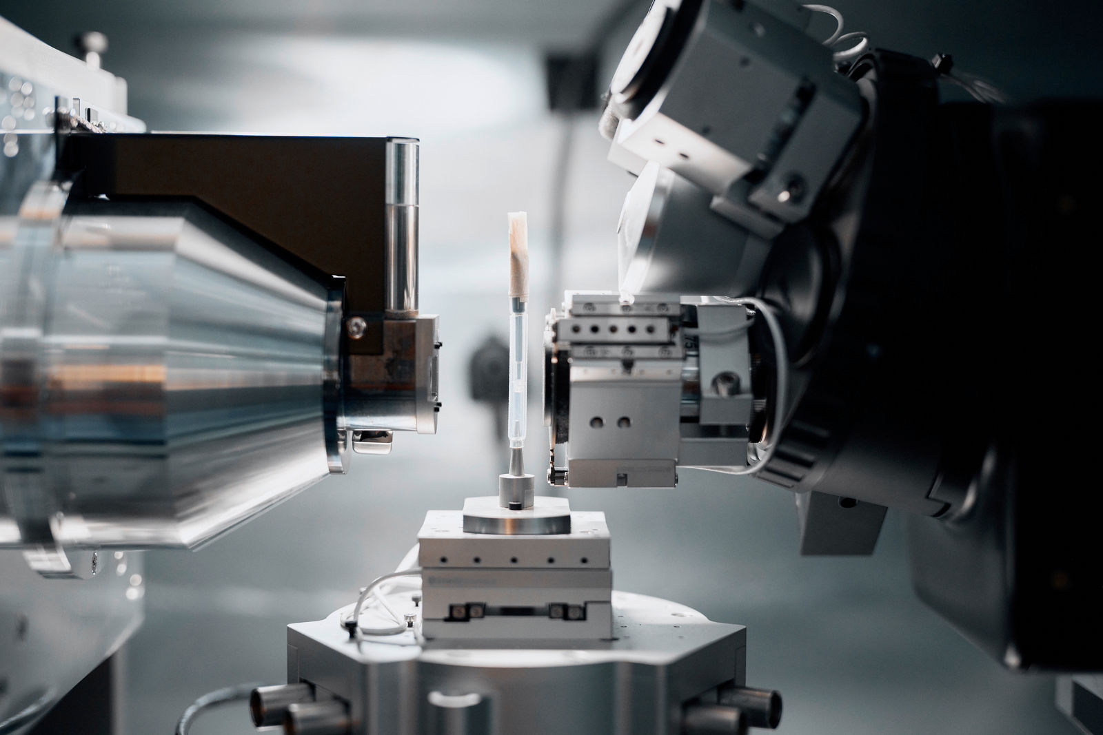 The image shows a high-precision scientific or industrial measurement and analysis device. At the center, there is a small sample held in place, surrounded by complex mechanical arms or optical sensors. This could be an X-ray diffraction (XRD) system or another high-resolution analytical device used in material science, battery research, or semiconductor manufacturing. The state-of-the-art equipment suggests a laboratory environment where material structures are analyzed at a microscopic or atomic level.