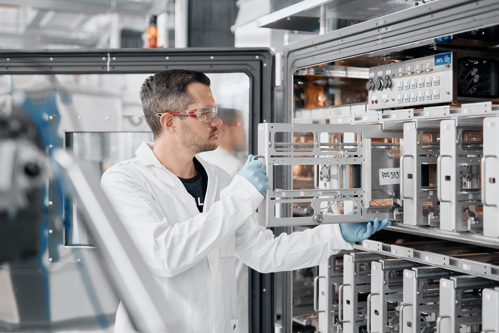 Das Bild zeigt einen Techniker oder Wissenschaftler in einem hochmodernen Labor oder einer Testeinrichtung. Er trägt Schutzkleidung, Handschuhe und eine Schutzbrille, was auf eine kontrollierte Umgebung für präzise Experimente oder Fertigungsprozesse hinweist. Die Anlage im Hintergrund scheint ein Testgerät für Batterien oder elektronische Bauteile zu sein, möglicherweise für die Automobil- oder Energiewirtschaft. Der Fachmann führt eine Prüfung oder eine Wartungsaufgabe durch, indem er eine Komponente in das System einsetzt oder entfernt.