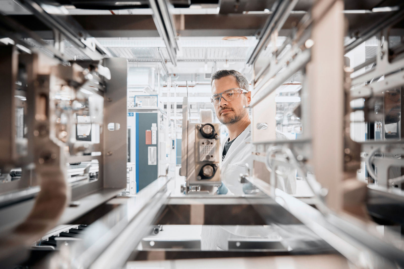 The image depicts a technician or engineer in a state-of-the-art laboratory or production facility. He is wearing safety glasses and a lab coat, indicating a controlled environment, possibly for research and development in battery technology or electromobility. The industrial equipment in the foreground suggests precision manufacturing or testing processes essential for high-tech components.