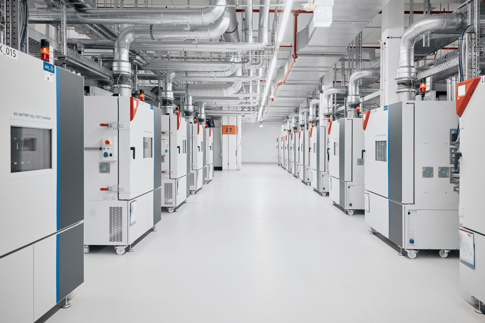 The image depicts a state-of-the-art laboratory or testing facility for battery cells. The hall is equipped with a series of test chambers, likely used for environmental or stress testing of batteries. The devices are connected to industrial ventilation systems, ensuring a controlled environment. The clean and organized surroundings indicate high safety and quality standards, which are essential for research and development in electromobility.