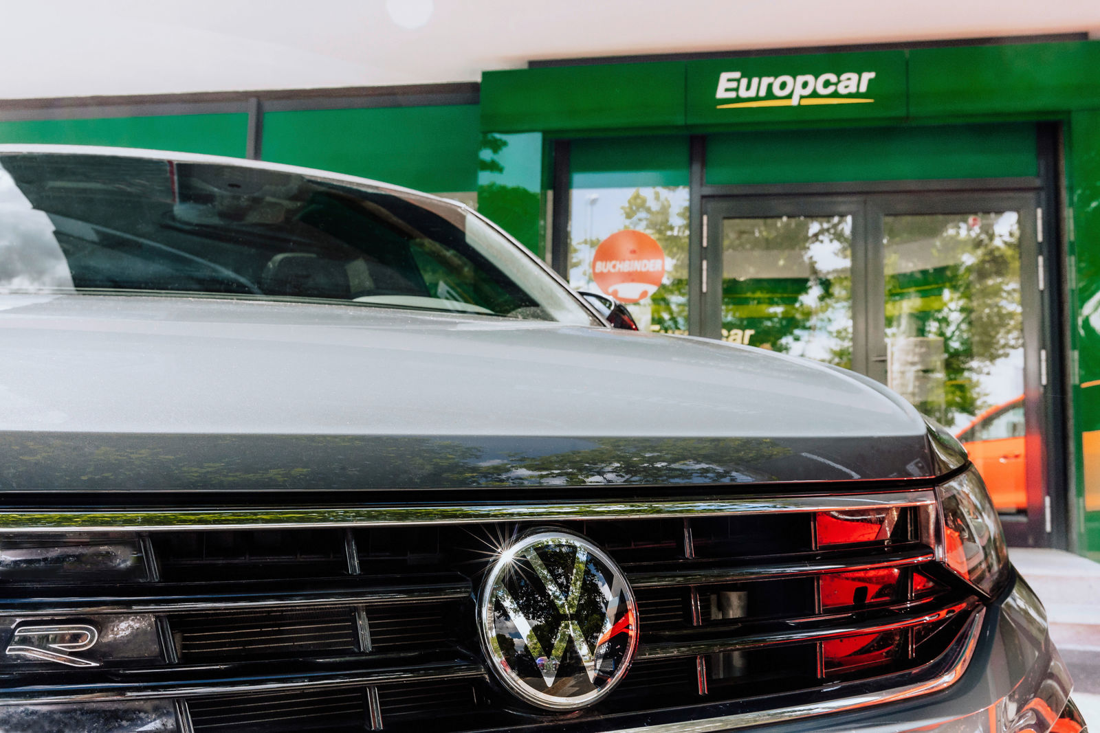 The image features a close-up of the front of a Volkswagen vehicle with a shiny VW emblem, parked in front of a Europcar rental location. The green Europcar signage is prominently displayed above the entrance, while another rental brand, Buchbinder, is reflected in the background. The scene suggests a partnership or rental service offering Volkswagen vehicles through Europcar.