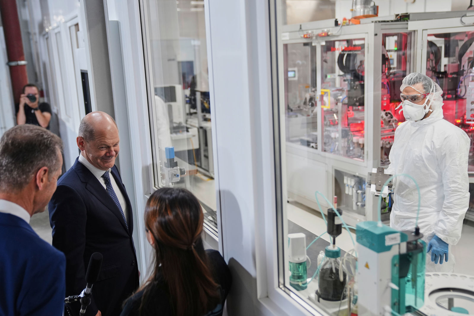 Chancellor Olaf Scholz visits a high-tech production facility and observes through a glass window a worker in protective clothing operating in a laboratory. In the background, a complex manufacturing line with machines and red lighting is visible.