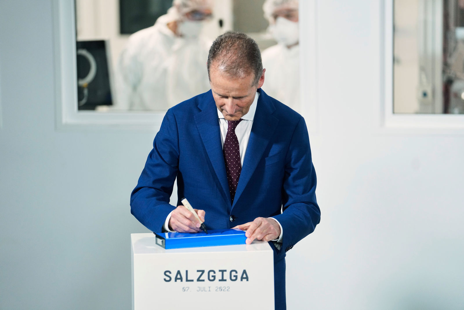 The image features a man in a blue suit signing on a tablet, which is placed on a white pedestal labeled “SALZGIGA 07. JULI 2022,” indicating an event related to a gigafactory in Salzgitter. In the background, individuals in protective suits suggest a high-tech production environment. The scene conveys the impression of an official milestone or a ceremonial signing as part of a major industrial project.
