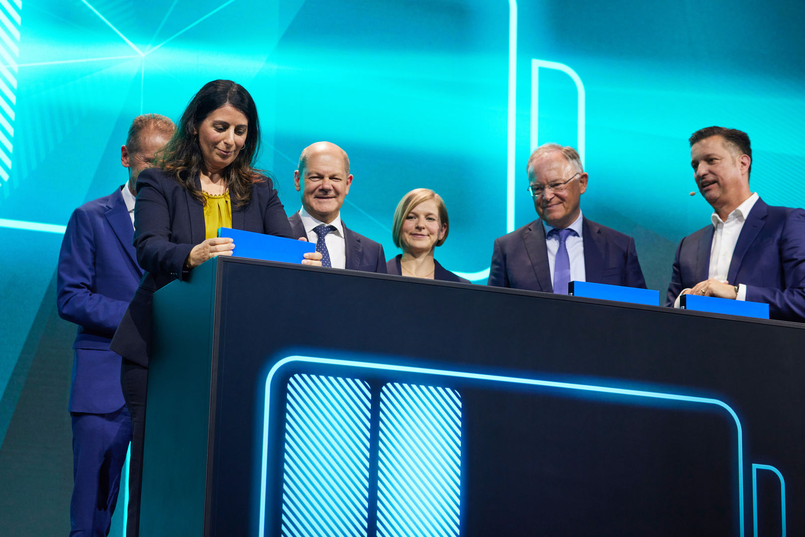 The image captures a ceremonial event on a stage with a futuristic, blue-lit backdrop. A woman dressed in dark clothing with a yellow blouse signs on a tablet while several individuals, including politicians and business representatives, look on with smiles. Among them, a recognizable political figure is present. The scene conveys the impression of a symbolic milestone, possibly related to a technology or industrial investment.